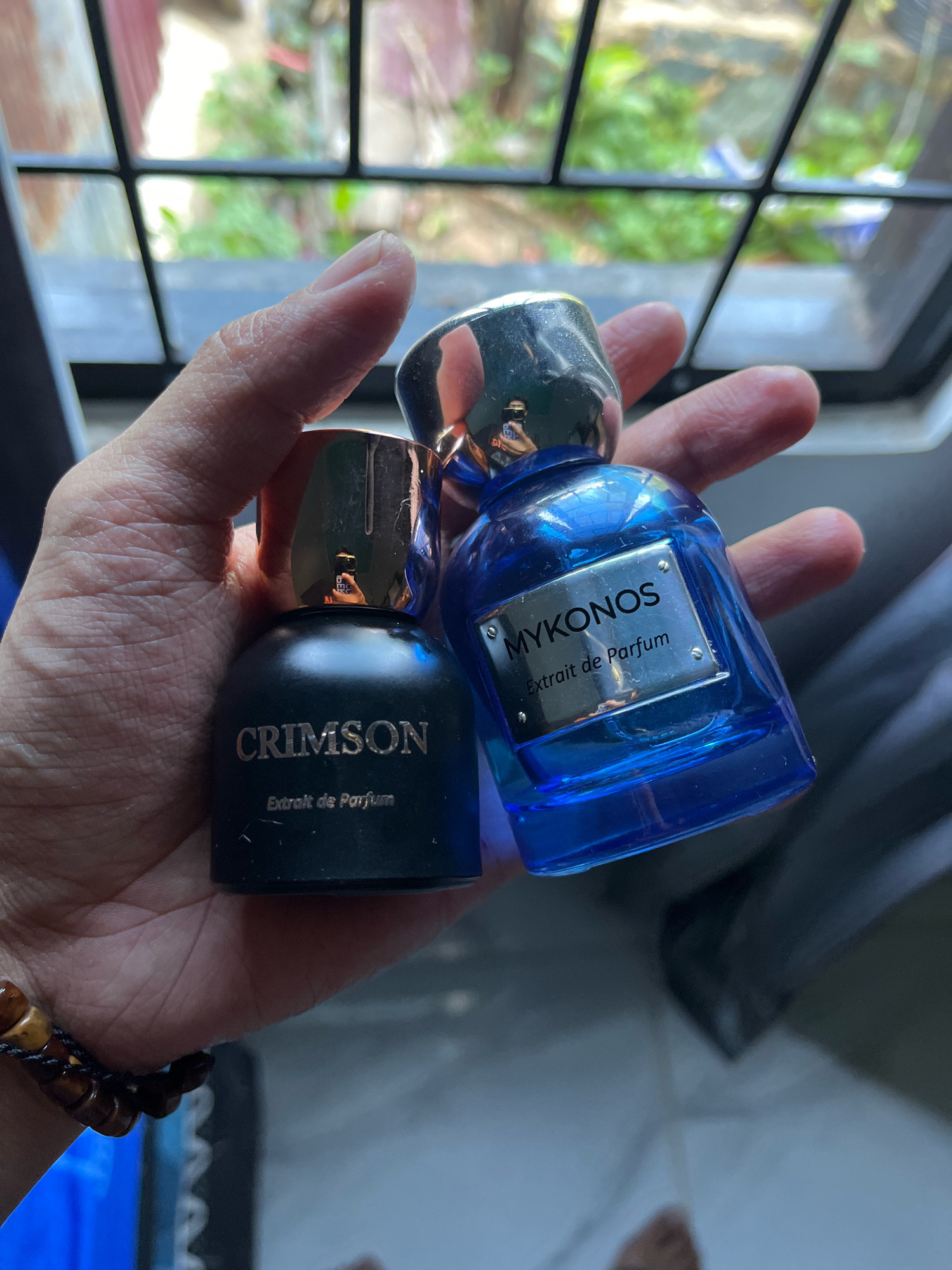 A close-up, high-angle shot captures a hand holding two perfume bottles in front of a window. The hand, with visible skin texture, gently cradles the bottles, one on the left and one on the right. The left bottle is matte black with a rose-gold cap, and the text "CRIMSON" is prominently displayed in white lettering, followed by "Extrait de Parfum" below. The right bottle is a vibrant blue glass, also with a rose-gold cap. A silver plate on the blue bottle reads "MYKONOS" in large white letters, with "Extrait de Parfum" written beneath it. A reflection of a person holding a phone is visible on the cap of the black bottle. The background is a window with a grid pattern, revealing greenery and parts of a structure outside, suggesting an outdoor or semi-outdoor setting. A beaded bracelet with brown wooden beads is visible on the wrist of the hand holding the bottles. The scene appears to be indoors, in Tungkaran Pangeran, Indonesia, near a window with a view of foliage and architectural elements.