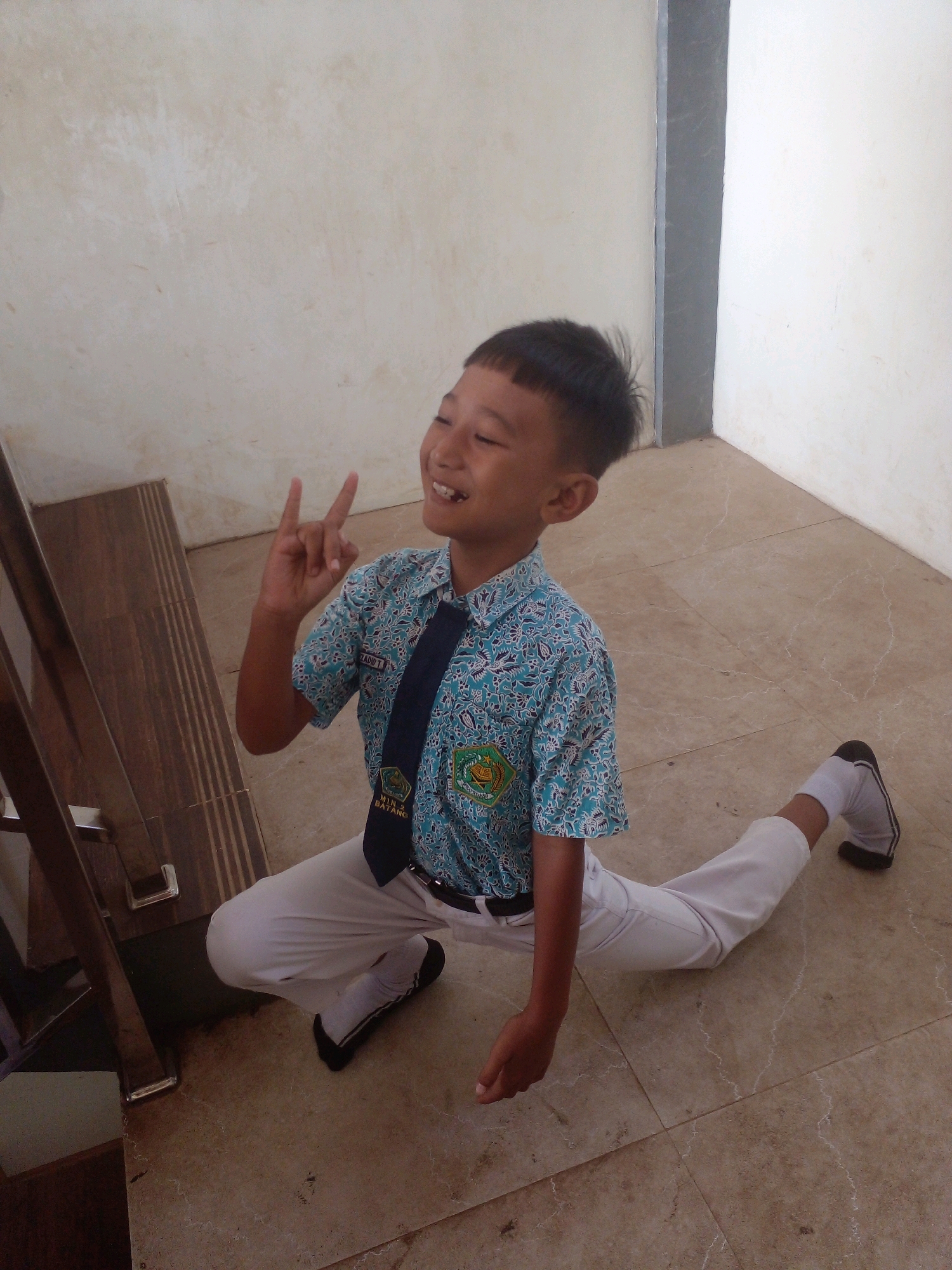 A young boy in a school uniform is shown in a split pose on a tiled floor. The boy's uniform consists of a blue patterned short-sleeved shirt, a dark blue tie, white pants, and black shoes with white socks. He is smiling and making a peace sign with his right hand. The environment appears to be indoors, possibly a school hallway or classroom, with light-colored walls and a wooden banister to the left. The image is taken from a slightly high angle. The text on his tie is partially visible and suggests the school is in Banyuputih, Indonesia.