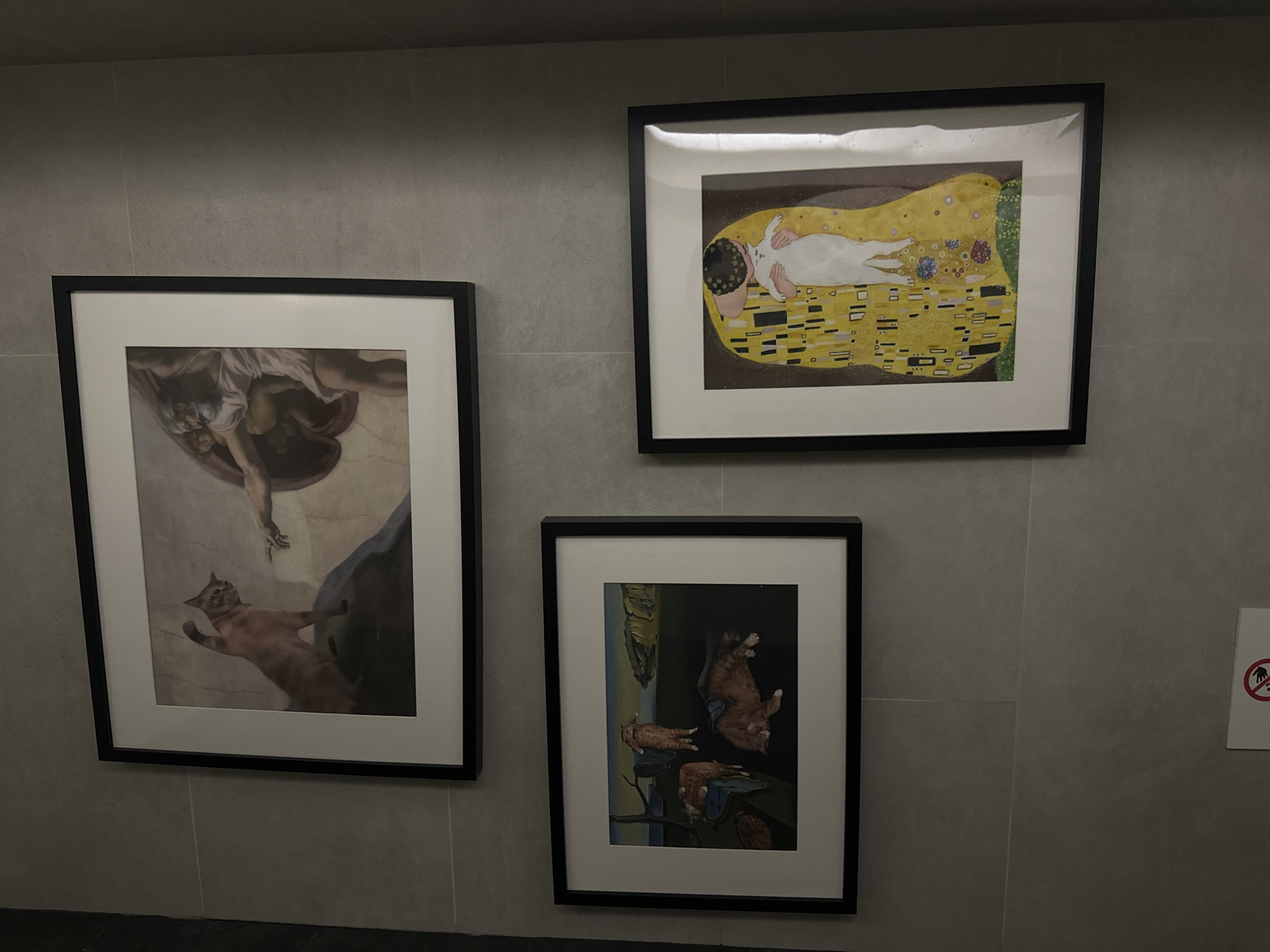 Three framed artworks are displayed on a plain, light gray tiled wall.
The top-right frame contains a parody of Gustav Klimt's "The Kiss," where a white cat replaces one of the human figures, lying on a patterned yellow blanket while being embraced. The background is dark brown.
The left frame features a parody of Michelangelo's "The Creation of Adam." A human hand extends from the upper part of the frame, reaching towards an orange cat below, mimicking the iconic pose. The cat appears to be in mid-air or suspended, with its paws extended.
The bottom-right frame shows a parody of Salvador Dalí's "The Persistence of Memory." Several orange cats are depicted in a surreal landscape, some appearing to "melt" or draped over objects like the iconic clocks in the original painting.
To the far right, on the wall, a small white sign with a red circle and a crossed-out hand symbol is visible, typically indicating "no touching" or "do not touch." The overall lighting is dim, and the scene appears to be indoors.