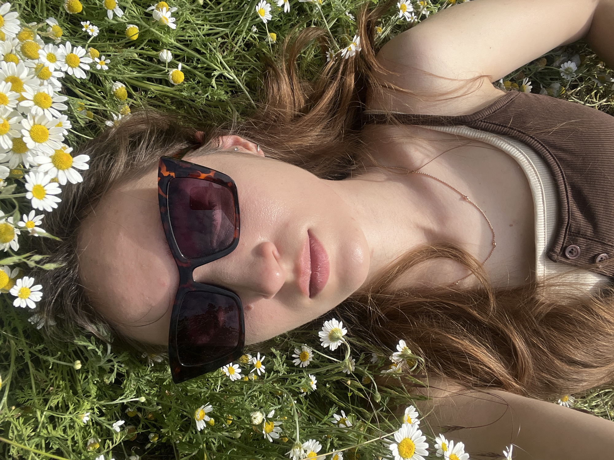 A woman with long, auburn hair lies on her back in a grassy field dotted with white and yellow daisies. She is wearing large, tortoiseshell sunglasses, a thin gold necklace, and a ribbed brown tank top over a beige undershirt. Her head is tilted back, and her face is illuminated by sunlight, casting a shadow from her sunglasses. The camera is positioned directly above her, looking down. The background is a soft blur of green grass and flowers, indicating an outdoor setting.