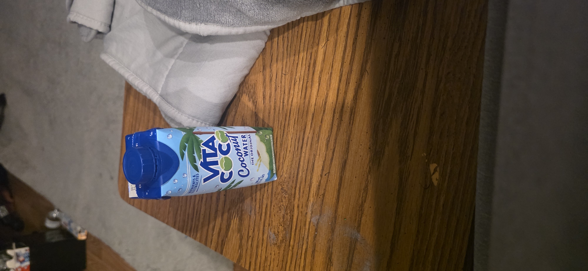 A carton of Vita Coco Coconut Water, 100% original, is standing upright on a wooden table. The carton is blue and white and features an image of a coconut and palm trees. The table is light brown with a wood grain texture. To the right of the carton, a folded gray blanket or towel is partially visible. In the background, a gray carpet is visible, along with some out-of-focus furniture. The scene appears to be indoors, likely in a residential setting in Kenner, United States of America.