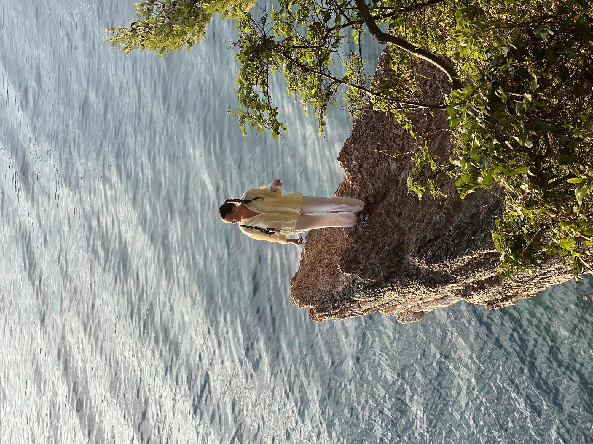 A woman stands on a rocky outcrop overlooking the Adriatic Sea in Kamenovo, Montenegro. The woman is wearing a yellow blouse and white pants, and has her hair in braids. She is looking out at the sea. The rocky outcrop is covered in vegetation, and the sea is a deep blue color. The sun is shining, and the water is calm.