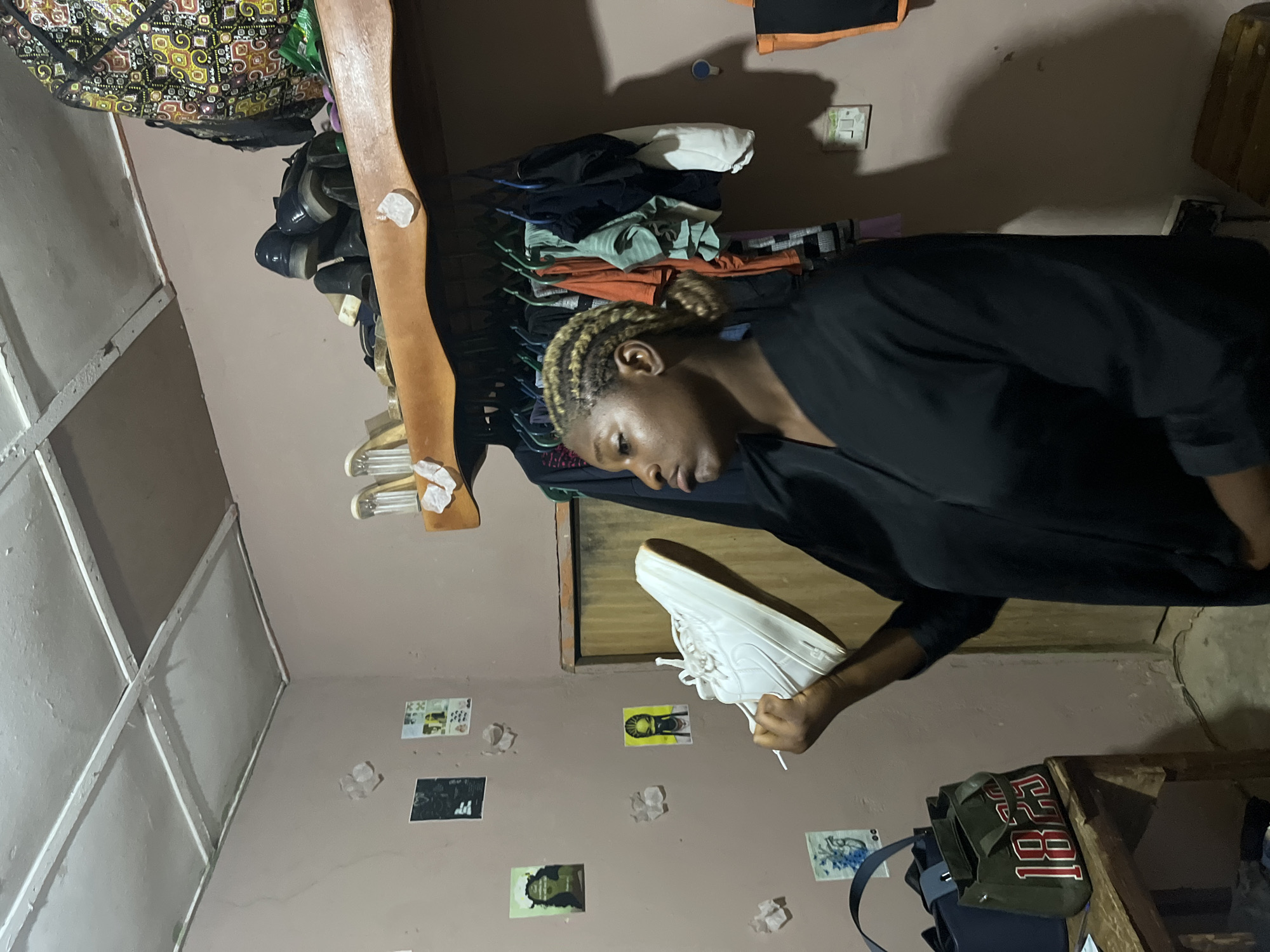 The image depicts an indoor scene, likely a room in Rafin Par, Nigeria. A young woman with braided blonde hair is standing and holding a white sneaker in her right hand, examining it. She is wearing a black garment. Behind her, the walls are painted a light brown and decorated with several small posters and what appear to be pieces of paper. A shelf above her head holds a collection of shoes, including black dress shoes and clear heeled sandals, as well as a patterned bag. Below the shelf, a rack of clothes hangs on hangers. To the left of the woman, a wooden table holds two handbags, one dark blue and another green with the numbers "1823" in red and white. The ceiling appears to be made of suspended panels.