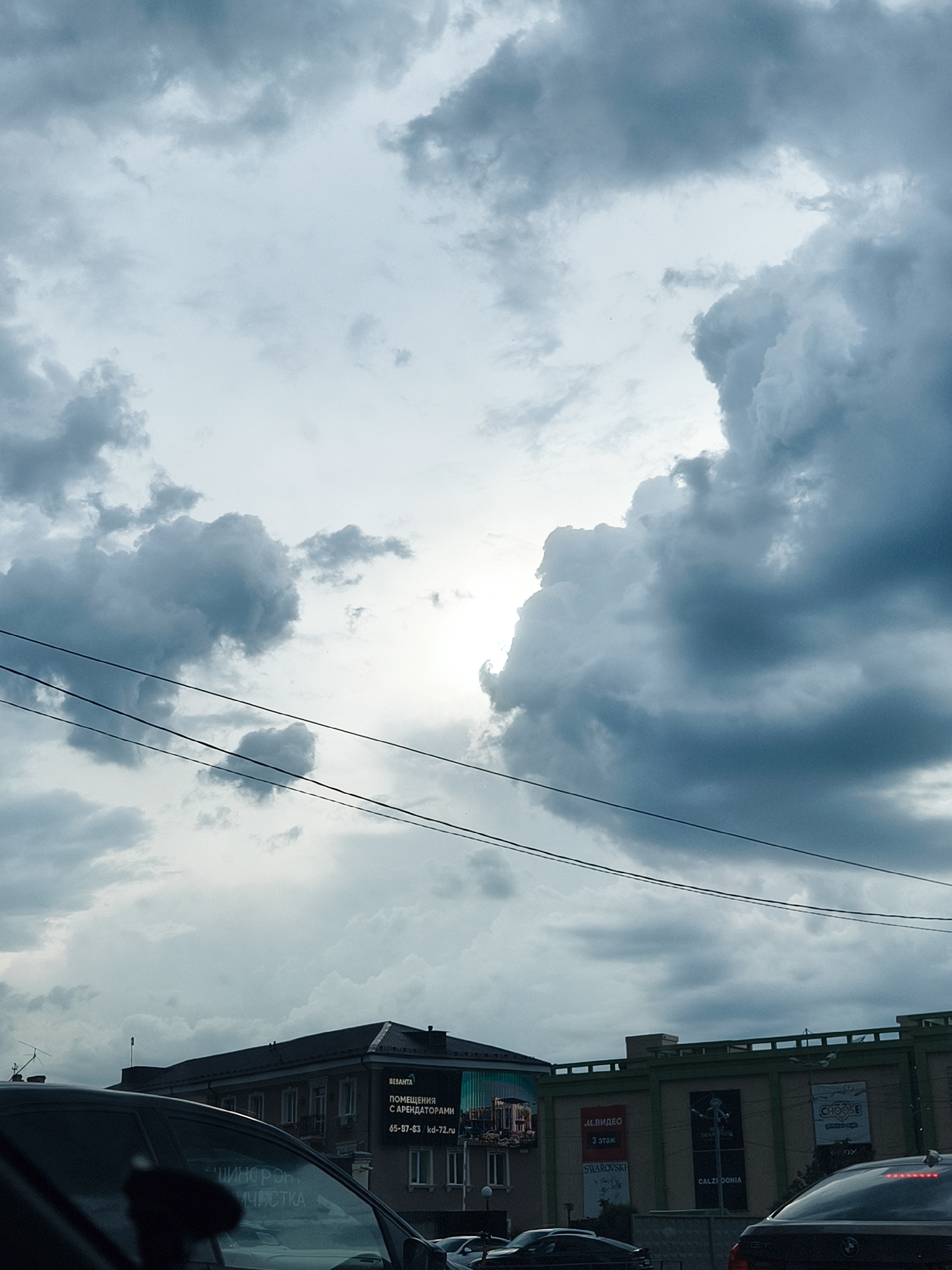 The image depicts a cloudy sky over a city street. The sky is filled with gray and white clouds, with a bright patch of light suggesting the sun is behind the clouds. Power lines stretch across the sky in the foreground.
Below the sky, buildings are visible, with advertisements and signs. A billboard for "BEBAHTA" advertises commercial spaces. Another sign advertises "M.VIDEO" on the 3rd floor. The text "SWAROVSKI" and "CALZEDONIA" can also be seen.
The lower portion of the image shows cars, likely stopped in traffic, with the rear of one car visible on the right.
The image appears to be taken from within a car.
Based on the advertisements and text visible, the image is taken in Tyumen, Russia.