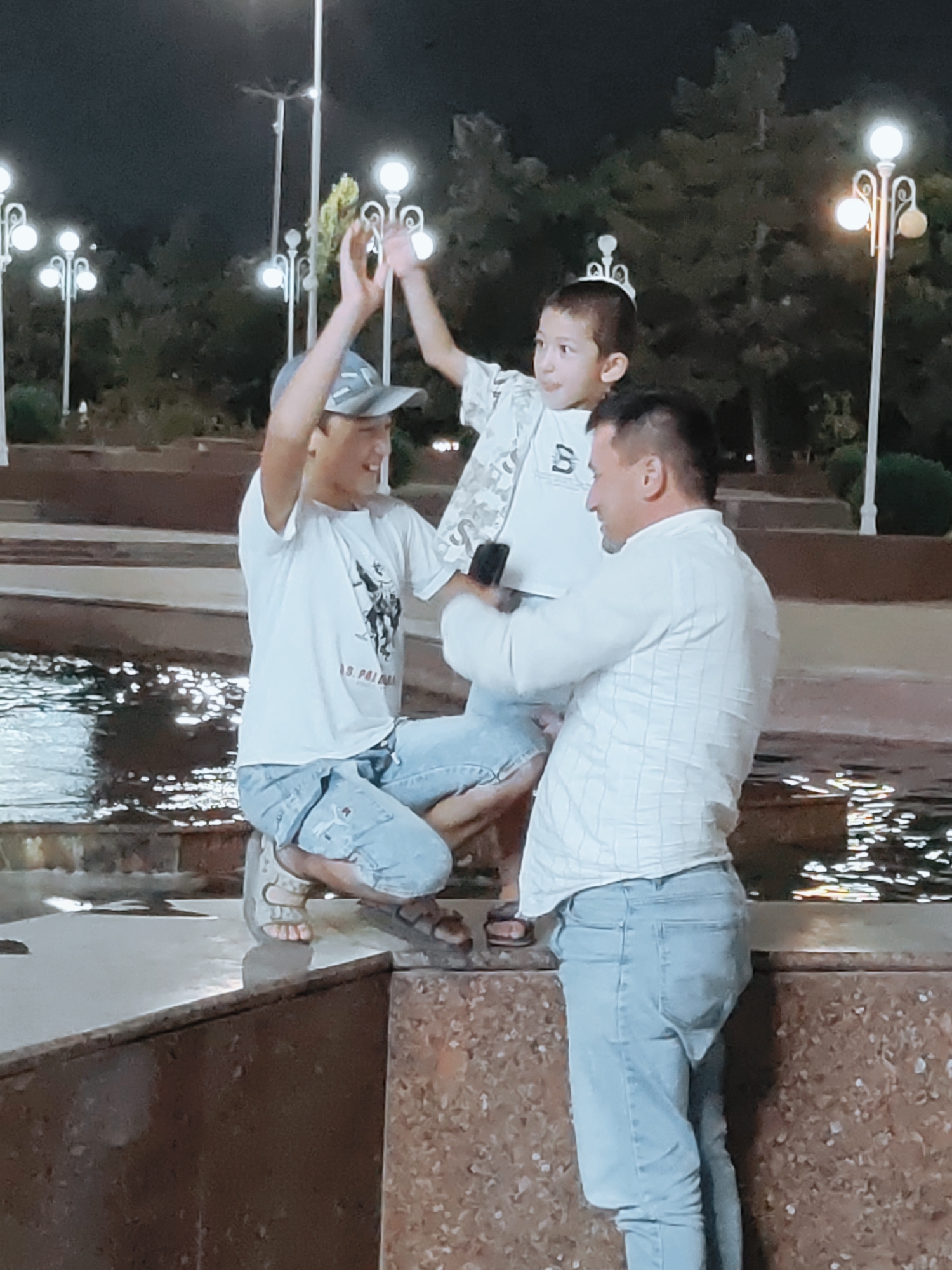 The image shows three individuals at night in a public park in Tashkent, Uzbekistan, likely near a water feature, given the reflections on the ground. In the foreground, a young boy with dark hair, wearing a light-colored t-shirt with a graphic print and a small crown-like accessory on his head, is being lifted by two other people. He is looking to his right and has his hands raised. To his left, a young man wearing a white t-shirt, a baseball cap, and light-colored jeans is partially visible from the waist down, crouched and looking up at the boy with his hands raised. To the right of the boy, an adult male, dressed in a white, checkered long-sleeved shirt and light-colored jeans, is holding a mobile phone in his right hand, seemingly taking a picture or recording a video of the boy. The background features illuminated streetlights and trees, typical of an urban park setting at night. The ground appears to be a combination of textured stone and a water surface reflecting light.