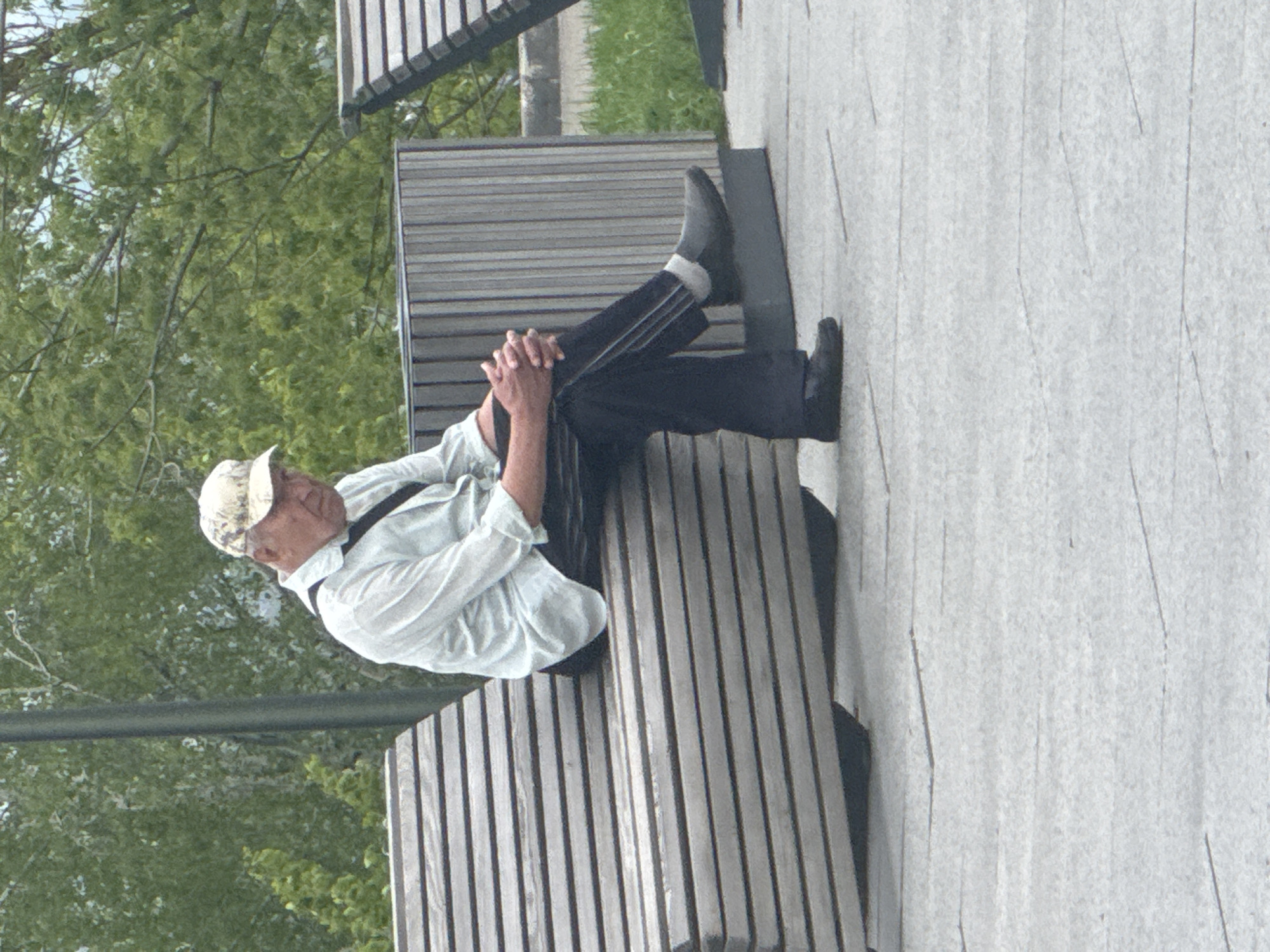 An elderly man in a light-colored shirt, dark pants, and a patterned cap is resting on a park bench. His legs are crossed and resting on the opposite end of the bench, with his hands clasped together near his knees. He is wearing dark shoes and light-colored socks. The bench is made of light-colored vertical slats. The bench is located on a paved surface, with green trees visible in the background. The location is in Nizhny Novgorod, Russia.
