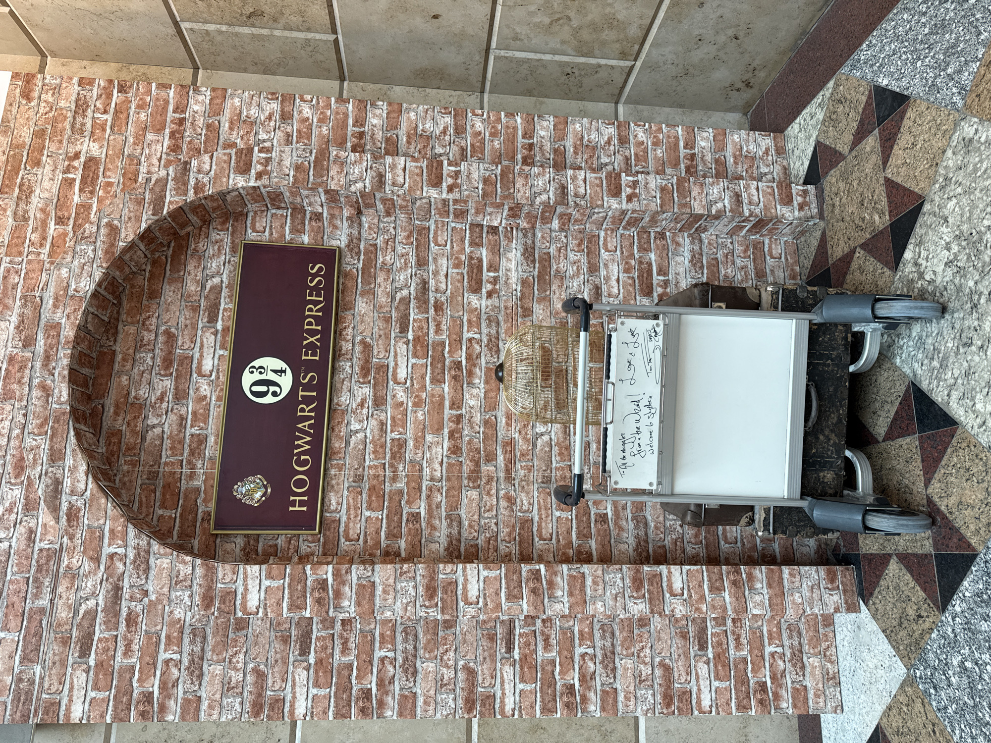 The image shows a re-creation of the Platform 9 3/4 scene from the Harry Potter franchise. A faux brick wall forms an archway. Inside the archway, a sign reads "Hogwarts Express" with the "9 3/4" symbol. In front of the wall is a luggage trolley with a birdcage and a piece of luggage on it. There is a whiteboard on the trolley with writing on it, including the phrase "Welcome to Slytherin." The location is most likely a theme park or exhibit in Moscow, Russia.