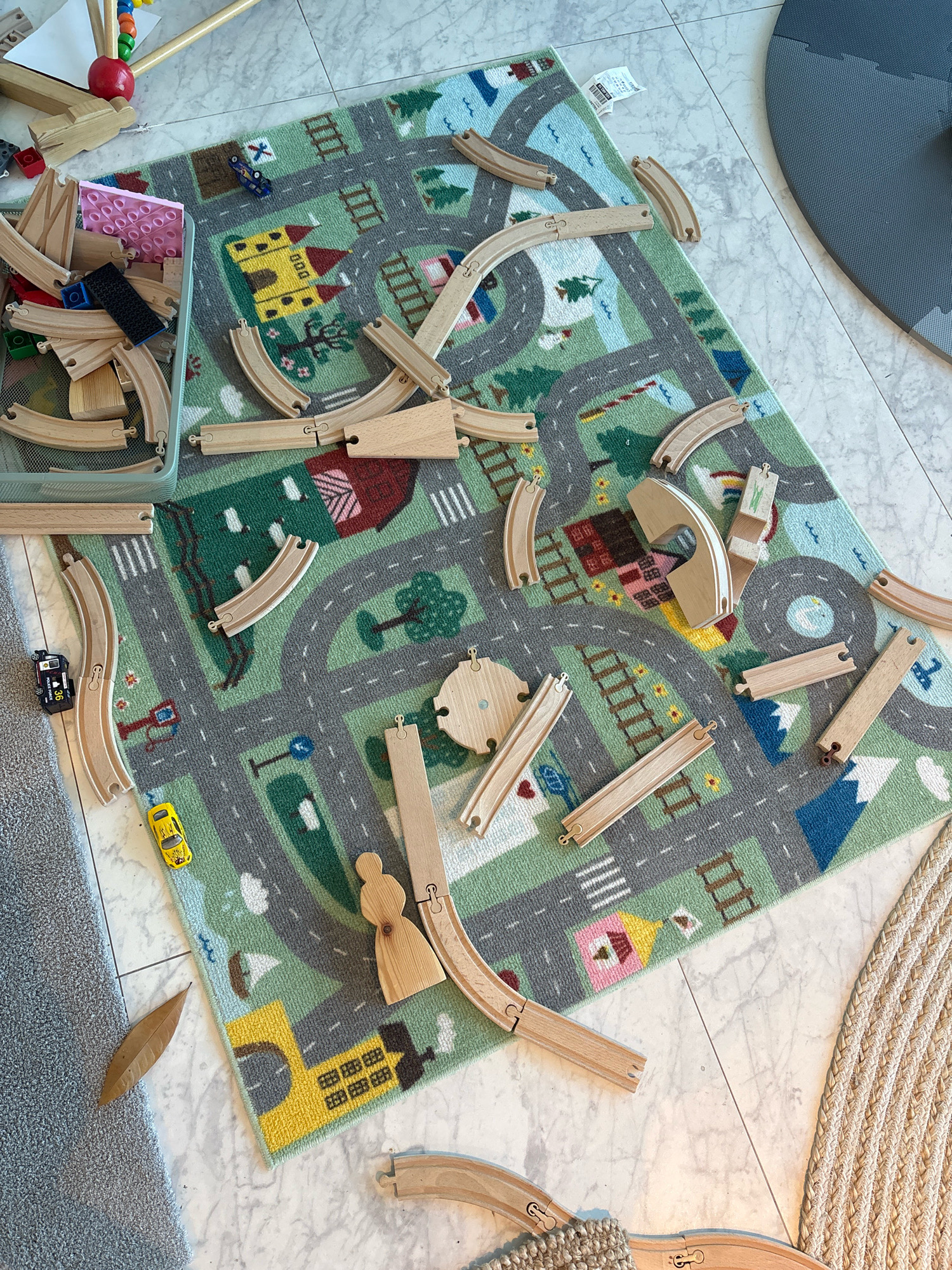 In this top-down, eye-level shot, a playroom floor in what appears to be a modern apartment in Reykjavik, Iceland is occupied by a colorful children's rug depicting a town with roads and buildings. Various wooden train track pieces are scattered across the rug, some connected to form partial tracks. Among the tracks, a small black toy police car and a yellow toy car are visible. To the left of the rug, a gray mesh bin is filled with more wooden train track pieces and some colorful building blocks. A single dried brown leaf rests on the floor near the bottom left corner of the image. The floor itself is tiled with a light gray and white marble pattern. A portion of a dark gray exercise mat is visible in the upper right corner. No people are present in the image.