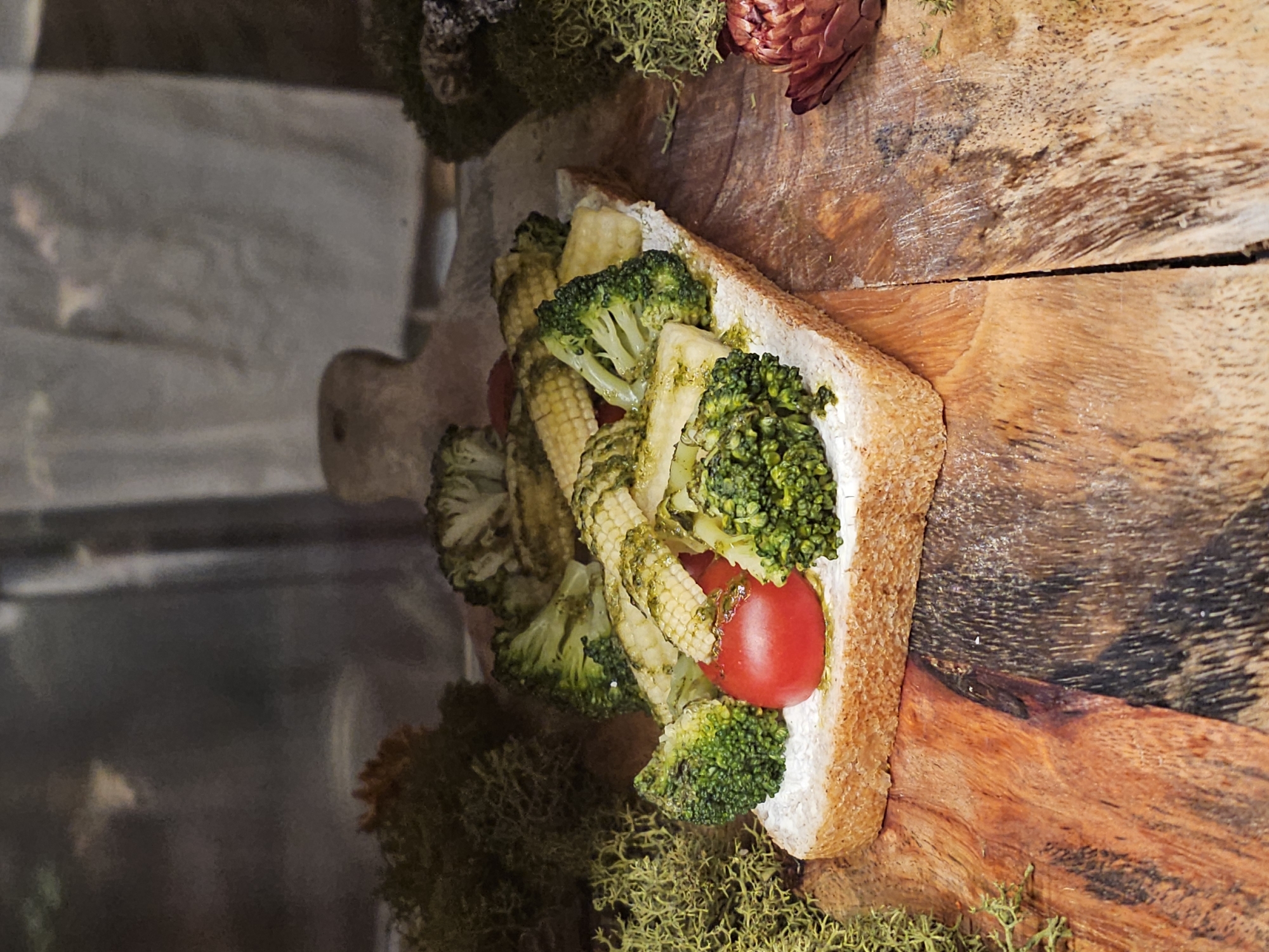 An overhead shot shows a piece of toast spread with white cheese and topped with broccoli, baby corn, and cherry tomatoes, all drizzled with a green sauce, placed on a wooden cutting board. The board is surrounded by moss and dried flowers, with a blurred background of a draped fabric. The scene is illuminated by soft light, creating a naturalistic aesthetic. There is no discernible location in Nemchinovo, Russia, nor any people or actions depicted.