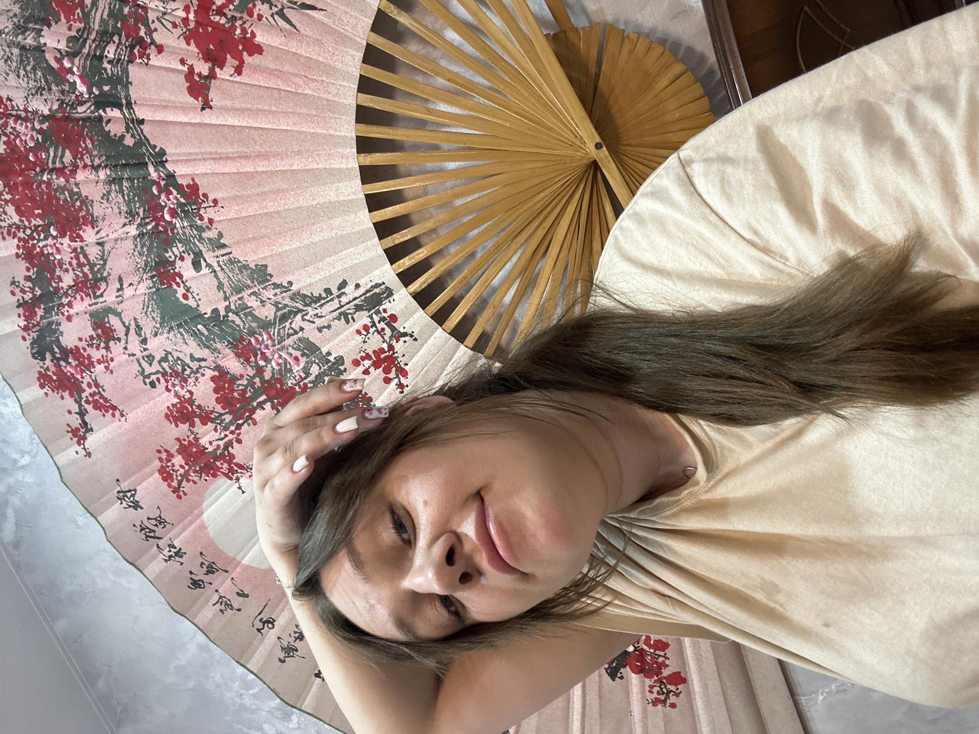 A woman lies on a surface covered with two Asian-style fans. One fan is pink with a floral design and Asian characters. The other is a plain, wooden fan. The woman is wearing a beige shirt and has long brown hair. Her right hand is resting on her head. The background is partially obscured, but appears to be indoors. There is no evidence in the image to suggest that the location is in Ольховка, Russia.