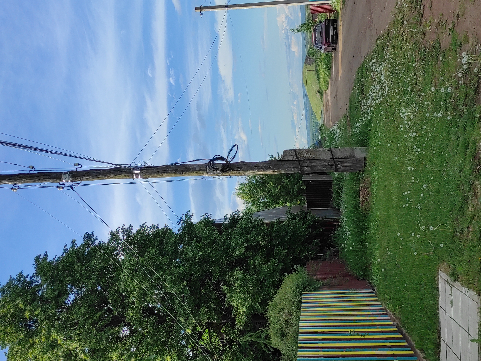 The image depicts a rural scene, likely in the vicinity of Olkhovka, Russia. A utility pole with power lines runs vertically through the left half of the image. The sky is blue with scattered clouds. To the right of the pole, the landscape slopes downwards, revealing a view of green hills. A dirt road runs along the top right edge of the image, with a dark-colored vehicle parked. Lush green vegetation and dandelions grow along the roadside. In the lower portion of the image, there's a section of a colorful striped fence or barrier. The scene is bright and appears to be taken during the daytime.