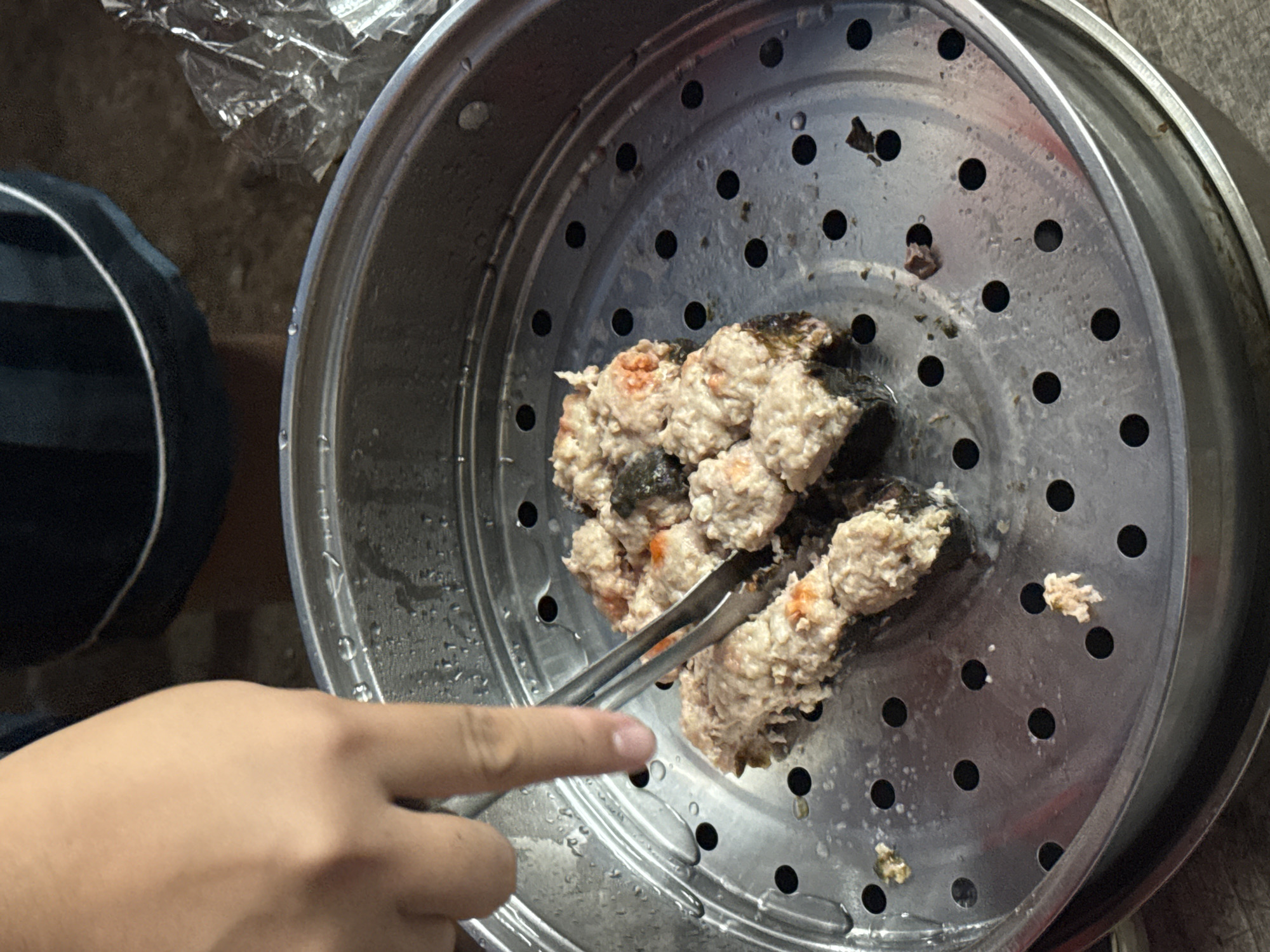 The image captures a close-up, top-down view of food items being handled inside a metal steamer. The primary subject is a human hand, light-skinned, seen from the wrist to the fingertips, holding a pair of metal tongs. The tongs are gripping several irregularly shaped, light-colored food pieces, which appear to be meat or fish balls, some with visible orange specks and wrapped in dark green/black seaweed or leafy material. These food items rest on a perforated metal steamer rack.
The steamer rack is round, silver-colored, and covered with condensation or water droplets, indicating it has recently been used for steaming. Beneath the rack, the base of a larger metal pot or steamer is visible. In the upper left background, some crumpled, clear plastic or foil is partially visible, and to the left, a dark blue/black object with a white trim is out of focus, possibly clothing or another container. The overall lighting suggests an indoor setting. There is no legible text visible within the frame. The activity depicts a person attending to freshly steamed food, likely preparing it for consumption. While the location context is provided as Davao City, Philippines, no specific visual cues within the image confirm this.
