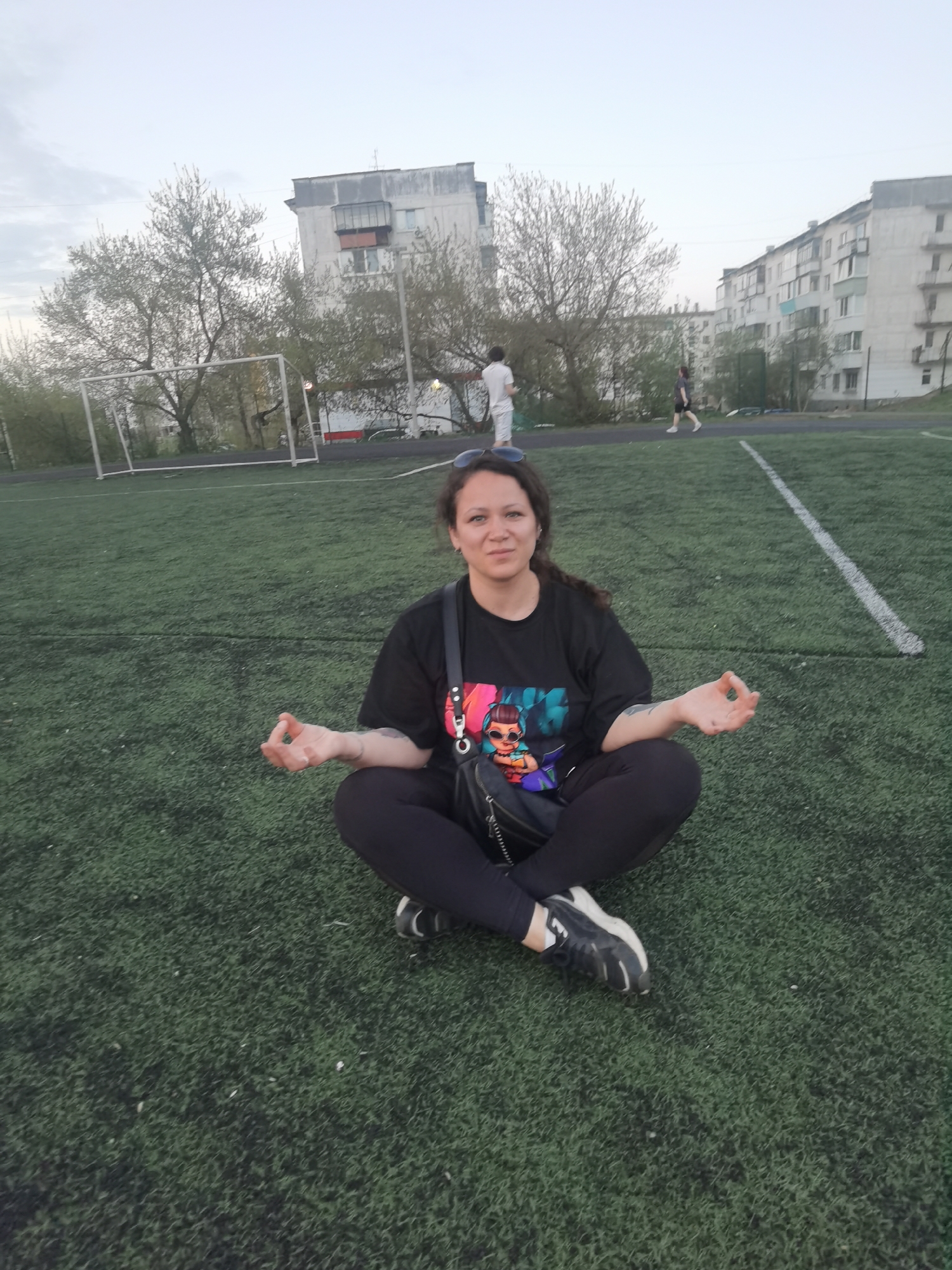 The image depicts a woman sitting cross-legged on an artificial turf field. She is wearing a black t-shirt with a colorful graphic print, black leggings, and black and white sneakers. She has her hands in a meditative mudra. A black crossbody bag is visible on her shoulder.
In the background, there is a soccer goal, buildings typical of Soviet-era architecture, and two people walking in the distance. The location appears to be a sports field in an urban environment. Based on the details and information available, the location is likely Chaikovsky, Russia.
