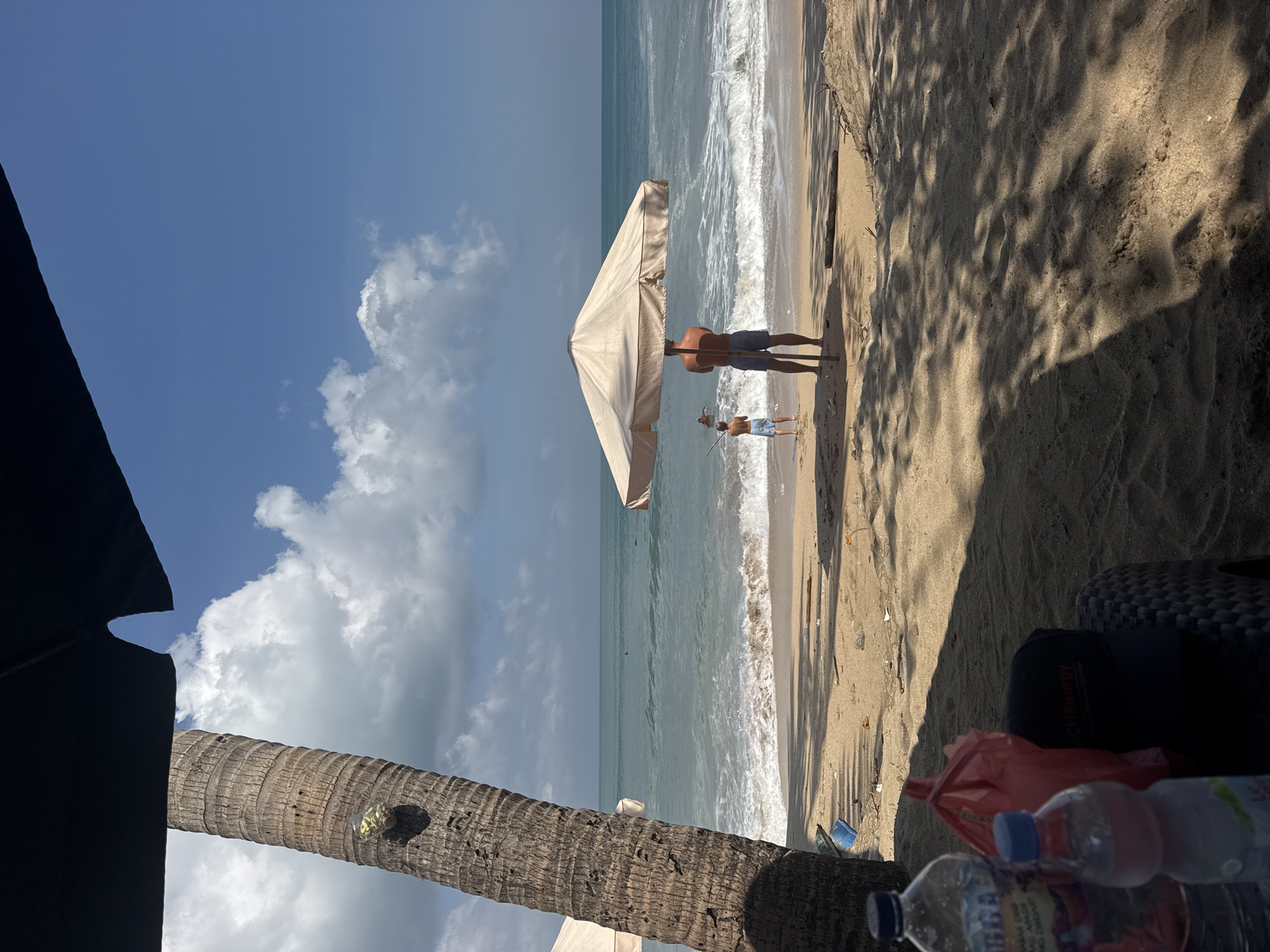 A beach scene in Kuta, Indonesia, features a sandy shore with gentle waves rolling in from the turquoise ocean under a bright blue sky with scattered white clouds. A prominent palm tree trunk stands on the left side of the frame, casting a shadow. In the mid-ground, a large beige beach umbrella shades two people: a man in shorts and a young boy in light blue shorts, both facing the ocean. The man stands close to the umbrella's pole, while the boy is a few feet away, looking towards the water. Further out in the ocean, several individuals are visible swimming. The foreground shows a table with two plastic water bottles and a red plastic bag, all resting on the sand. The beach appears somewhat littered with small pieces of debris.