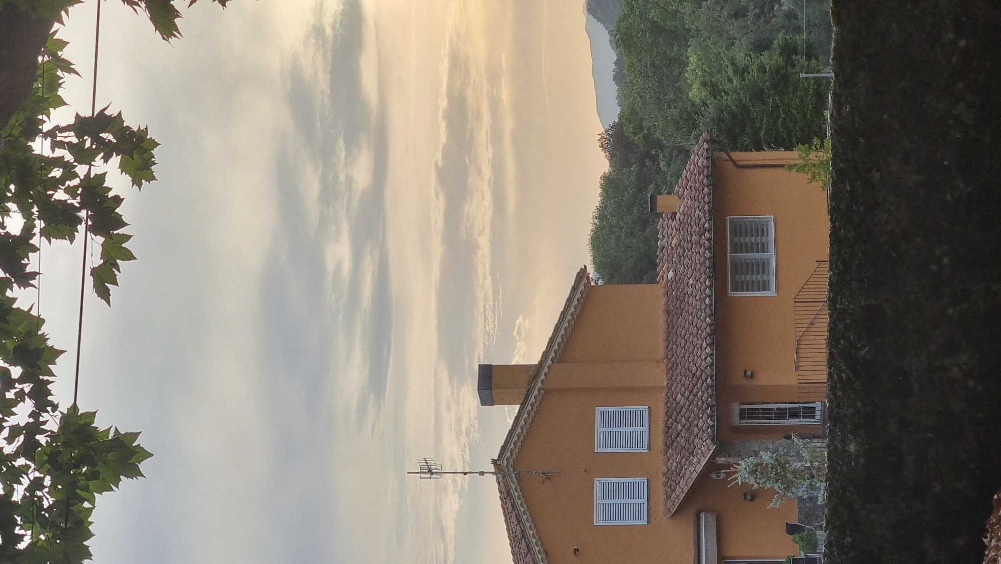 The image shows a two-story orange house with a red tile roof, located in Sant Joan Les Fonts, Spain. The house has white-shuttered windows on both levels and a chimney. An antenna is visible on the roof. In the background, there is a dense forest and rolling hills under a cloudy sky with hints of sunset. A dark, textured wall is in the foreground, obscuring the lower part of the house and yard. There are no people or animals visible in the image.