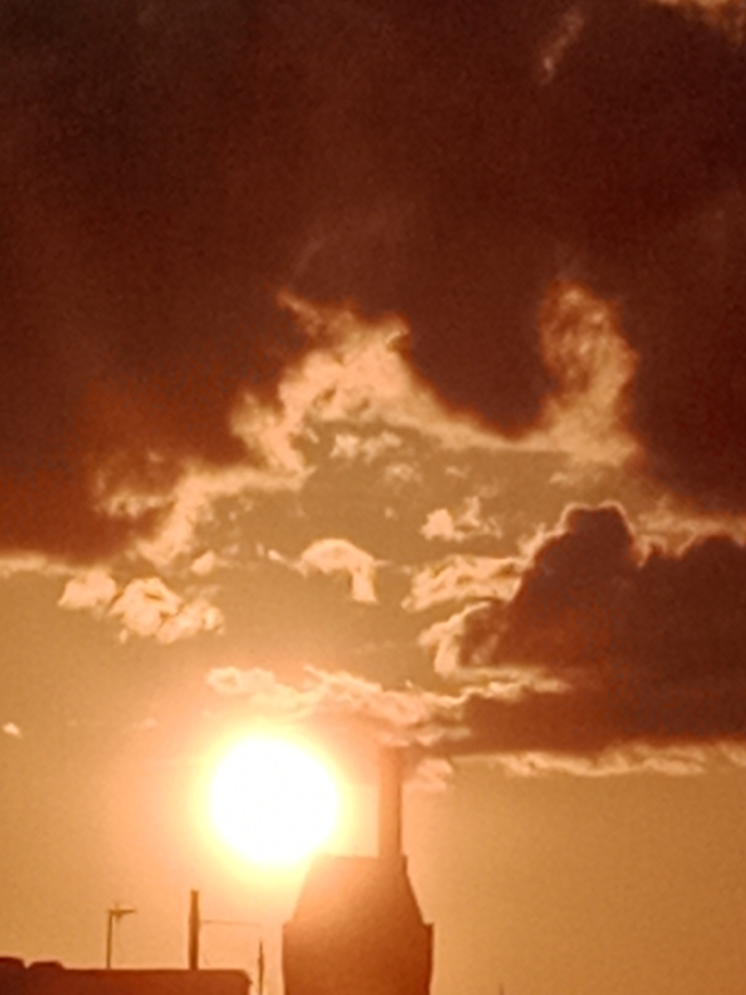 A sunset casts an orange glow over Bydgoszcz, Poland, illuminating dark clouds and silhouetting buildings and utility poles in the foreground. The sun, a bright, blinding disc, is partially obscured by a building's shape. The sky is a dramatic mix of dark, ominous clouds and lighter, illuminated ones, creating a stark contrast. The image captures a moment of natural beauty with an urban backdrop.