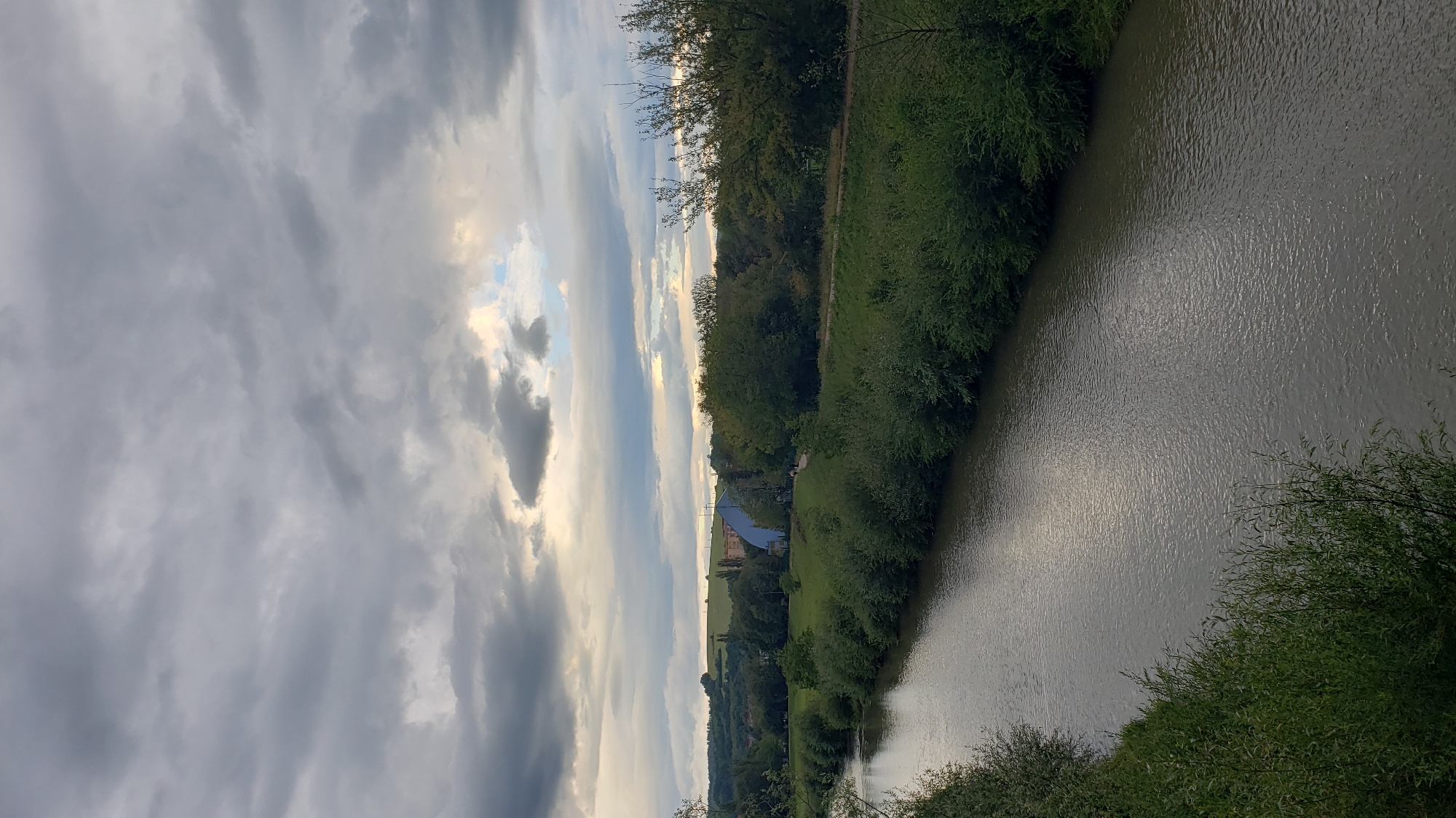 The image depicts a river flowing through a landscape near the city of Prešov, Slovakia. The river occupies the foreground, with its murky, rippling surface reflecting the overcast sky. Dense green foliage lines both banks of the river. In the midground, a verdant expanse of grass is visible, dotted with trees and a distinctive light blue, modern-looking building with a curved roof. Beyond this, rolling hills covered in grass and trees stretch towards the horizon. The sky is dramatically filled with a mix of dark, heavy clouds and lighter patches, suggesting an impending or recent rain shower. The overall scene conveys a sense of nature and semi-urban development under a moody sky.