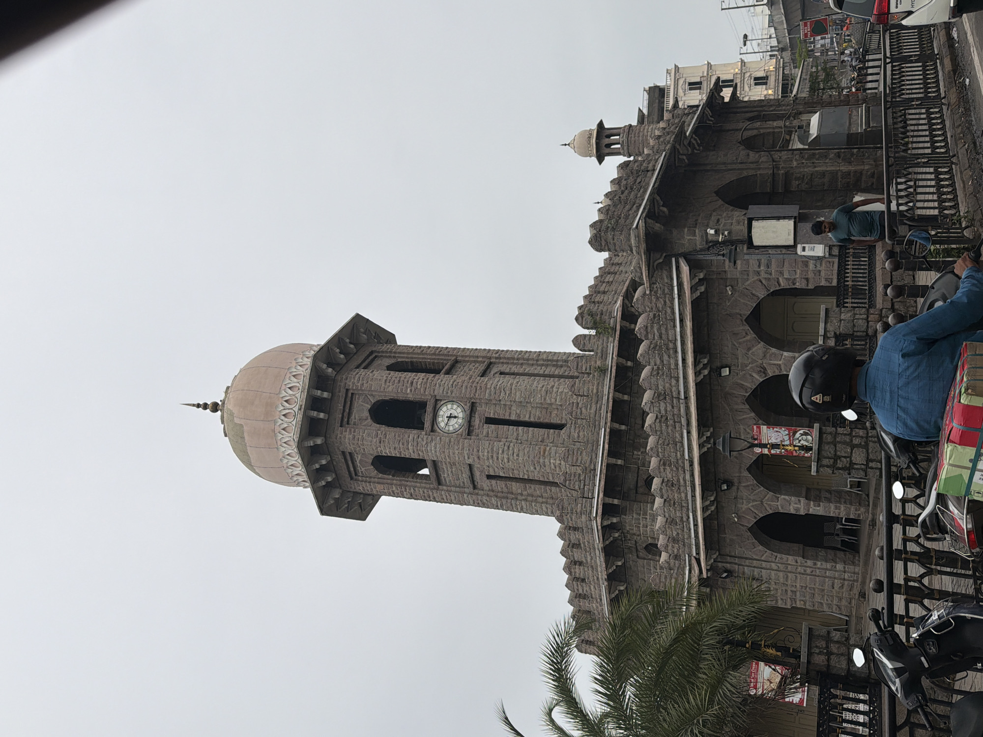 The image shows the iconic Clock Tower at Charminar in Hyderabad, India. The overcast sky suggests it's a cloudy day. The stone tower, topped with a pale pink dome, stands prominently. Below the dome, there is a clock face visible on one side of the tower. The tower is part of a larger stone structure with arched entrances and crenellated battlements. A palm tree is visible to the left of the tower. In the foreground, several parked scooters are visible, with a person in a blue shirt and helmet on one of them. Another person in a green t-shirt is standing near the arched entrances of the building. The scene depicts a busy street environment with traffic and pedestrians.