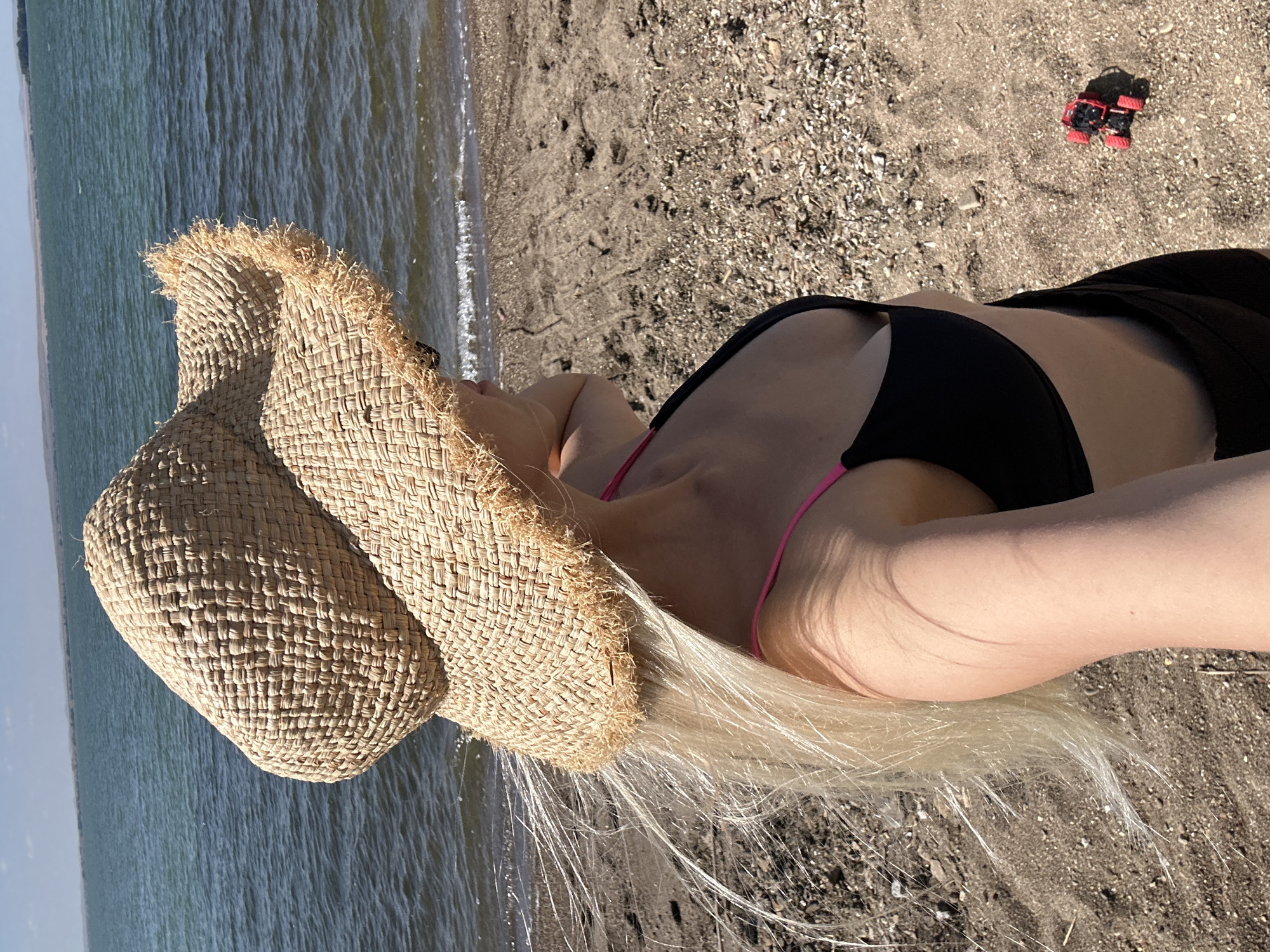An outdoor, eye-level, medium shot shows a woman from the chest up, facing right, standing on a sandy beach with a body of water and a distant horizon behind her. She is wearing a wide-brimmed, woven straw hat, a black bikini top with pink straps, and black shorts. Her long, blonde hair is partially visible beneath the hat and is blowing in the wind. The sand is light brown and scattered with small pebbles. The water is a deep blue-green with small waves. A tiny red toy car is visible on the sand to the right of the woman. The sky is clear and light blue. The location is not specified as Energetik, Russia.