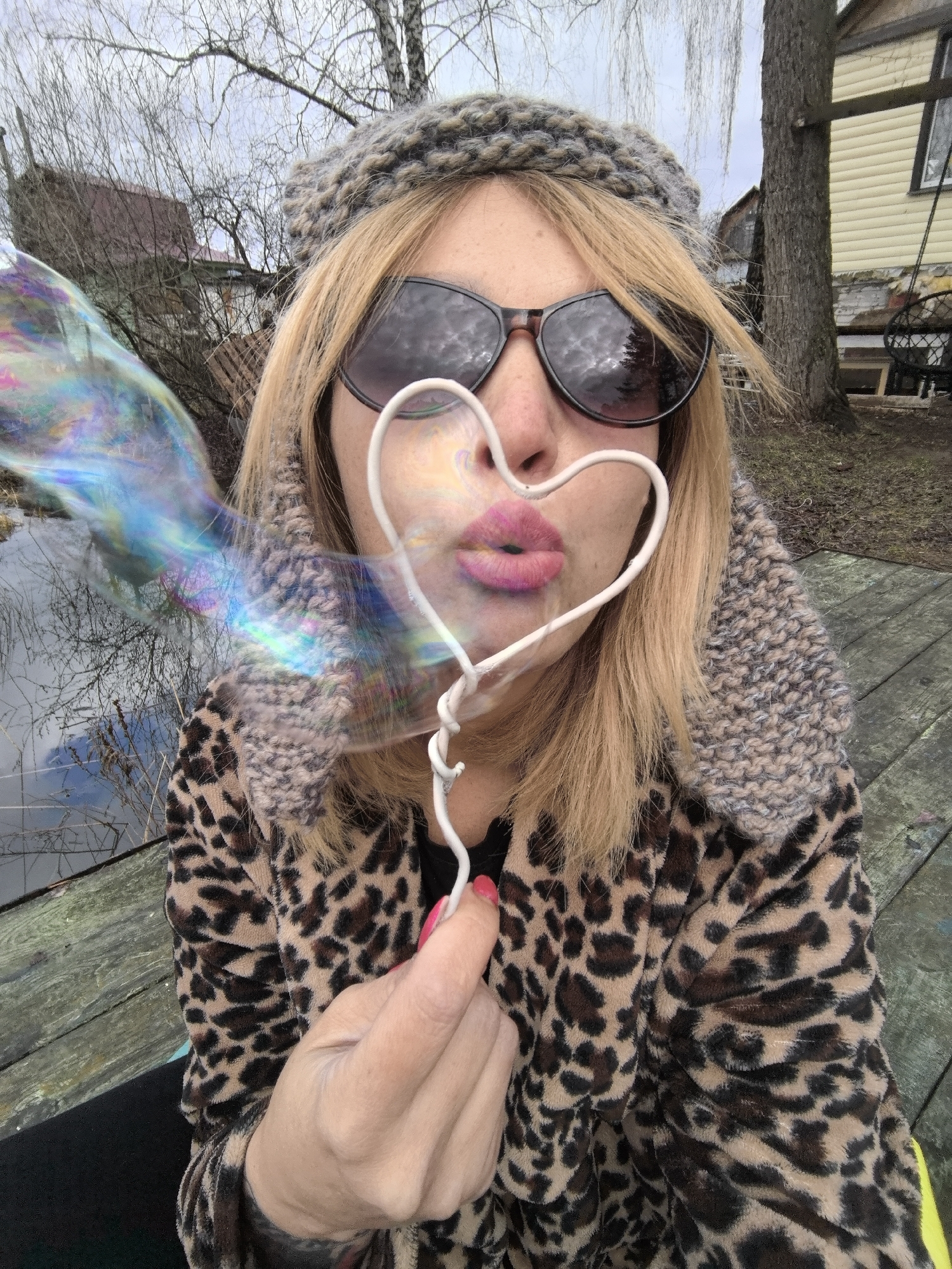 An outdoor, close-up shot captures a woman blowing bubbles in Noviy, Russia. She is positioned centrally, her head and upper torso filling most of the frame. The woman has blonde hair, wears dark sunglasses, and a textured, ear-flapped knit hat, along with a cozy leopard print jacket. Her lips are puckered around a white, heart-shaped bubble wand, and a large, elongated iridescent bubble emerges from it, displaying vibrant hues of blue, pink, and green. This bubble partially obscures the left side of her face and the background.
The setting suggests late winter or early spring, characterized by bare trees and an overcast sky. The woman is seated on a weathered wooden structure, likely a dock or deck, next to a still body of water that reflects the bare branches of trees. In the background, a light-yellow sided building with a darker roof is visible through the trees. The overall mood of the image is playful and lighthearted. There is no visible text in the image.