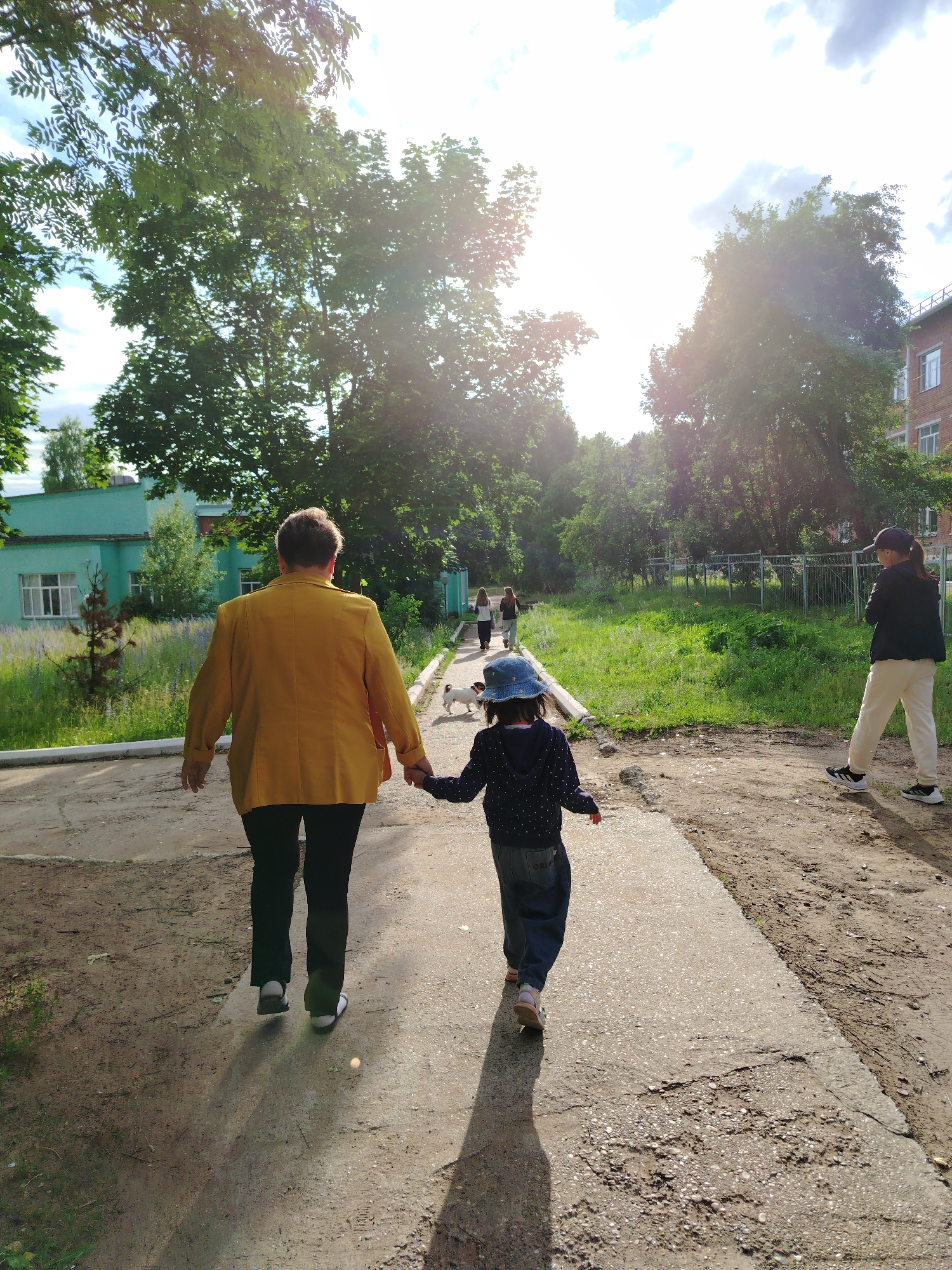 In Noviy, Russia, on a sunny day, a woman in a yellow jacket and a young girl in a blue hat are walking hand-in-hand down a paved path. They are walking away from the camera, with the woman on the left and the girl on the right. The girl is wearing a dark blue polka dot shirt and dark pants, and the woman is wearing black pants.
Behind them, further down the path, are three other people and two small white dogs. To the left of the path, there's a long, low green building with white-trimmed windows. To the right, there's a chain-link fence and a brick building. The scene is surrounded by lush green trees and grass, suggesting a park or a residential area with ample greenery. The sun is shining brightly, casting long shadows on the path, indicating it is likely late afternoon. The overall atmosphere appears peaceful and relaxed.