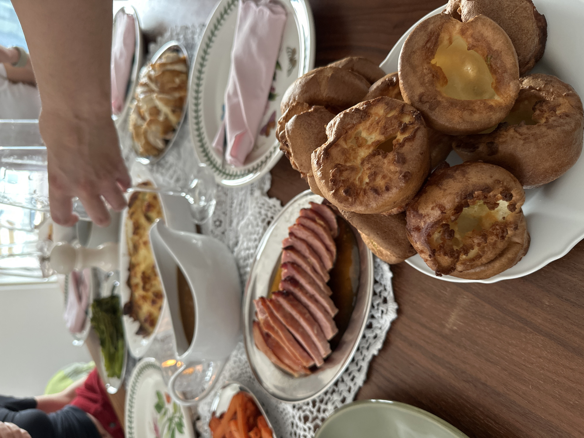 The image depicts a dining table laden with food, likely in a private residence. The table is covered with a white lace tablecloth. On the table, there are several dishes: a platter of Yorkshire puddings, a plate of sliced ham, a gravy boat, a dish of asparagus, a plate of carrots, and several other dishes with unidentified food items. A person's hand is reaching towards the food. There is also a stack of plates nearby. The environment suggests a dining room setting.