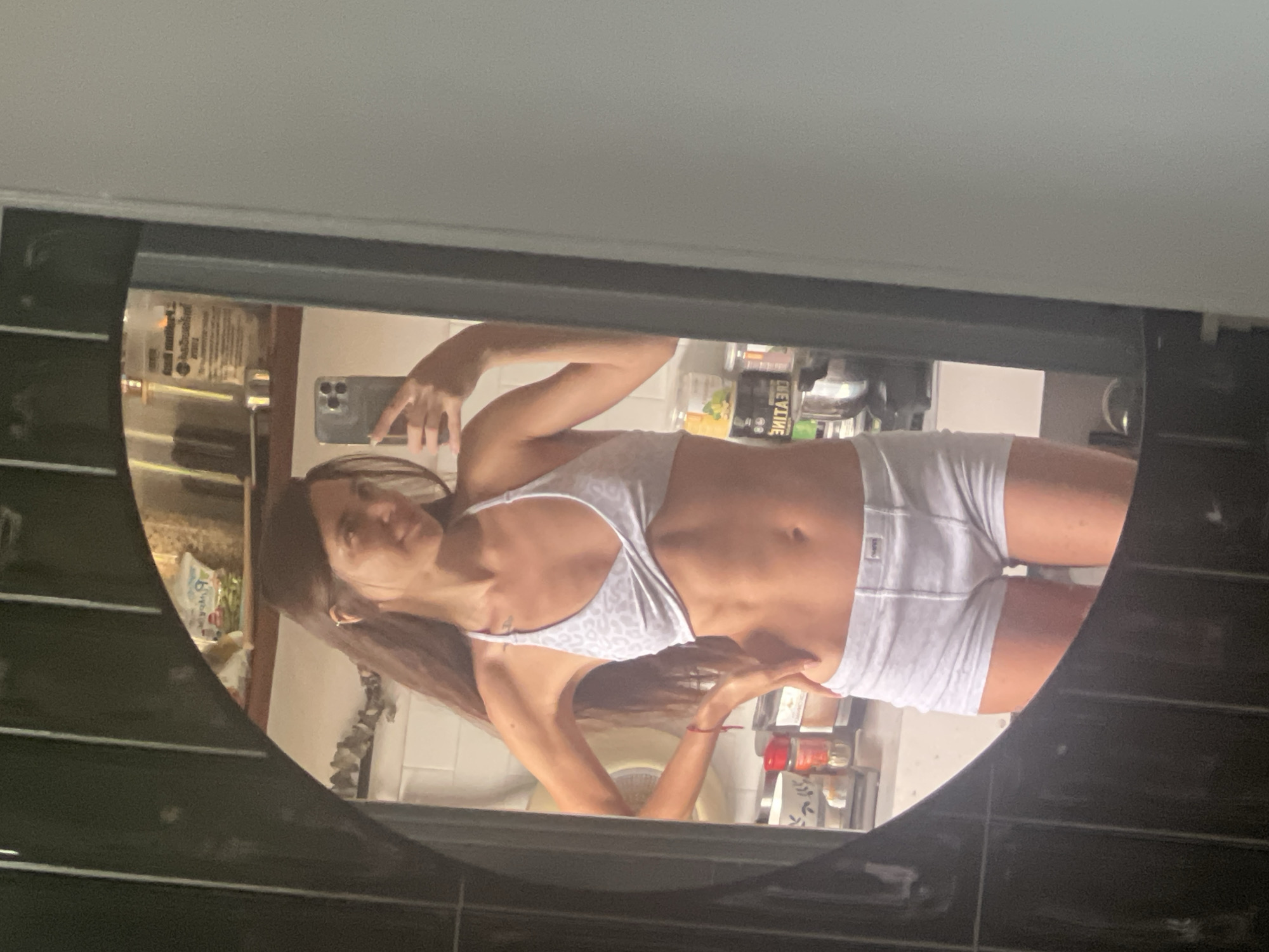 In an indoor setting, a woman with long brown hair is captured in a mirror selfie. She is wearing a white leopard-print sports bra and white boxer briefs. Her body is toned, and she is holding her left hand on her hip while her right hand holds a smartphone. The background, reflected in the mirror, shows a kitchen counter with various items, including jars and containers. The lighting suggests it is daytime.