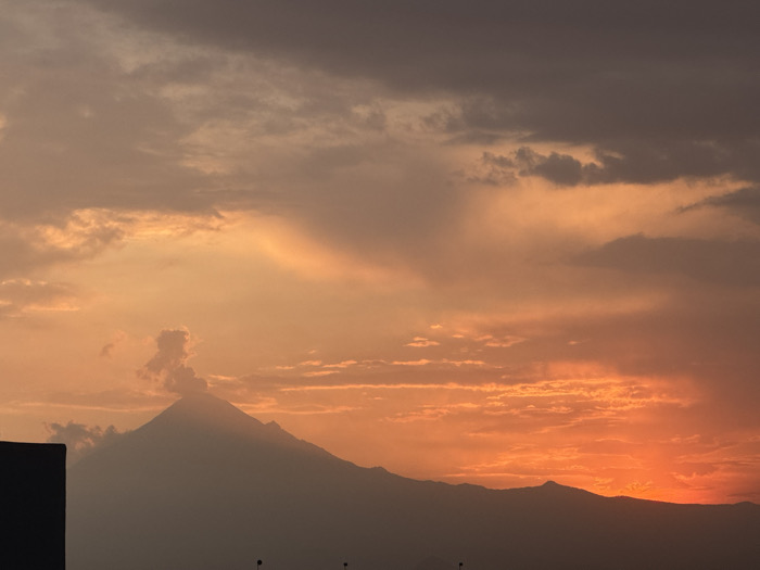 The image captures a twilight scene over San Francisco Ocotlán (Ocotlán), Mexico, featuring a mountainous landscape under a vibrant sunset sky. The sky displays a gradient of colors, transitioning from intense orange and red along the horizon on the right, through softer orange and peach tones, to dusky gray-purple hues towards the top and left. Scattered clouds are visible across the sky, illuminated from below. A prominent, dark-silhouetted stratovolcano dominates the mid-ground, positioned slightly left of center, from which a distinct plume of smoke or ash rises vertically from its summit. This volcanic peak is part of a larger, dark mountain range extending across the horizon. In the lower-left foreground, a dark, angular structure, likely a building edge, is partially visible. No individuals or discernible activities are present.