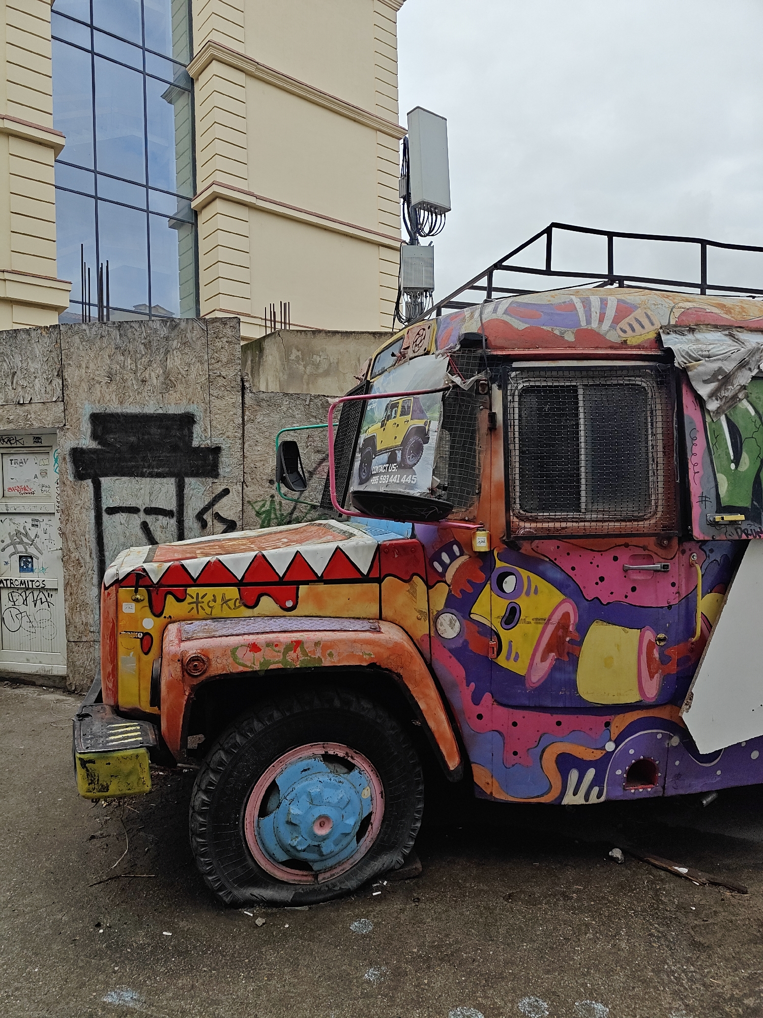 The image depicts a vibrant, graffiti-covered truck parked on an urban street in Tbilisi, Georgia, under an overcast sky. The truck, possibly a repurposed utility vehicle, is adorned with psychedelic, abstract art featuring a dynamic mix of bright colors like orange, purple, pink, yellow, and green. Patterns, including white and red zigzags, decorate the hood, which also bears the stylized graffiti "Keko."
The front left tire of the truck is completely flat, suggesting it has been stationary for some time. A black metal roof rack is mounted on top. On the driver's side window, a poster displays an image of a yellow Jeep alongside the text "CONTACT US:" and a Georgian phone number, "+995 593 441 445."
The truck is positioned next to a wooden barrier or fence, which is also heavily tagged with various graffiti including "TRAV," "ATROMITOS," and partially visible "DRUN." In the background, a modern building with a beige facade and large, reflective glass windows rises above the street, contrasting with the truck's colorful, weathered appearance. A cellular antenna array is mounted on a pole near this building. The ground is an asphalt surface with scattered debris, contributing to an overall urban, somewhat neglected yet artistically vibrant atmosphere.