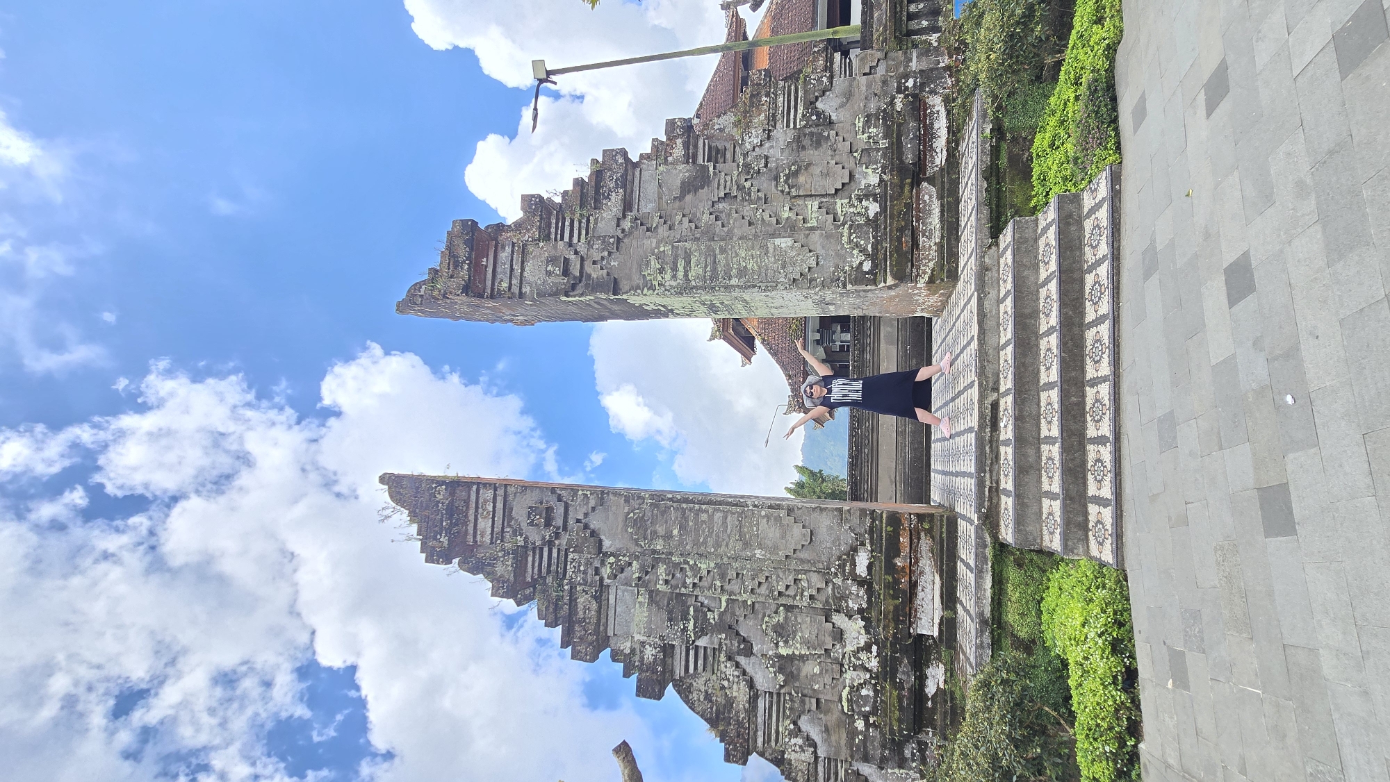 A vertical, wide shot captures a woman posing at the Handara Gate, a famous landmark in Baturiti, Indonesia. She stands on the stairs leading through the gate, arms outstretched. She is wearing a black dress with white horizontal stripes and a white hat. The gate itself is made of gray stone with intricate carvings and is partially covered in moss. The sky above is blue with scattered white clouds. To the right of the gate are green plants and a gray stone pathway. In the background, a red-tiled roof is visible, as well as distant mountains. A light pole extends into the sky.