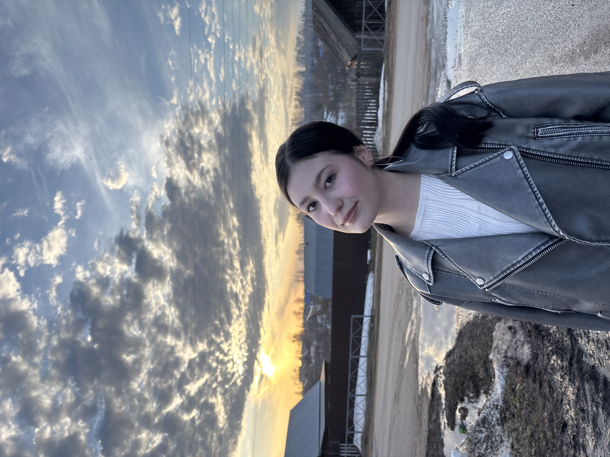 A young woman stands outdoors, wearing a grey leather jacket over a white top. The background reveals a landscape with clouds at sunset, a dirt road, and buildings, possibly in the city of Фоки, Russia. The ground shows patches of snow.