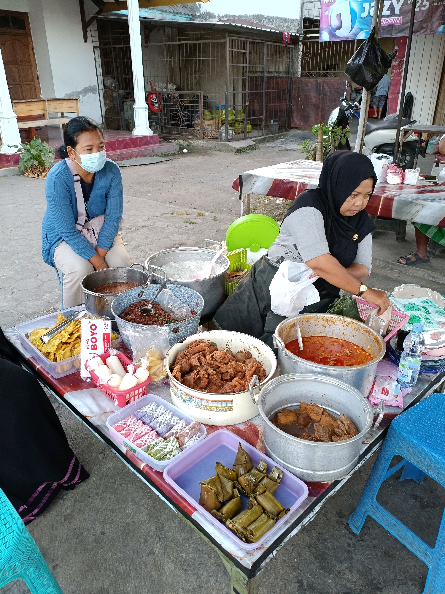 In Maguwoharjo, Indonesia, a street food vendor is seen tending to her stall. A woman wearing a blue cardigan and a face mask sits on a low stool to the left, facing away from the camera. To her right, a woman wearing a black hijab and grey shirt is actively serving food from large metal pots. She is holding a plastic bag and appears to be interacting with a customer or preparing an order. The stall itself is covered with a red, white, and blue patterned tablecloth and displays a variety of food items. These include fried items in a clear plastic container, small white cylindrical items in a pink basket, colorful rolled snacks in a clear rectangular container, and rice cakes wrapped in banana leaves in a purple tray. Several large metal pots contain cooked dishes, some with ladles. Bottles of water and other packaged goods are also visible on the table. In the background, a motorcycle is parked, and various structures suggest a typical street scene with buildings and outdoor spaces. The ground is paved with concrete. A bright blue plastic stool is positioned to the right of the stall.