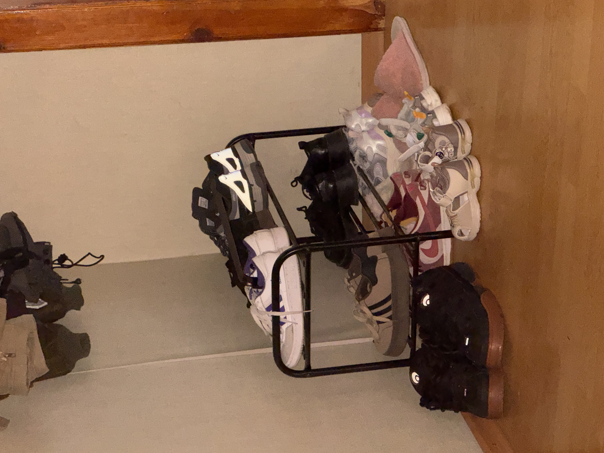 The image depicts an indoor scene, likely a closet or entryway, featuring a multi-tiered shoe rack overloaded with various footwear. On the top shelf, two pairs of black and white athletic shoes are visible. The second shelf holds a pair of white and purple sneakers. Below that, a pair of tan and dark blue athletic shoes are present. The bottom shelf and the floor area are crowded with more shoes, including black loafers, colorful sneakers, and a pair of pink slippers. Some clothing items, possibly jackets, are draped over the top of the shoe rack. The flooring is a light brown wood laminate, and the walls are a neutral beige. The lighting is dim, suggesting an indoor environment, possibly in the evening or a poorly lit room. There are no people visible in the image. The scene is consistent with a domestic storage space. The provided location of Яромінскі сельскі Савет, Belarus, does not appear to be directly represented or inferable from the visual content of the image itself.