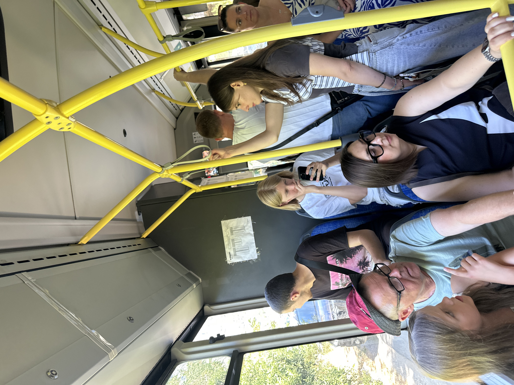 The image depicts the interior of a bus in Prčanj, Montenegro, with several passengers seated and standing. In the foreground, a young girl with blonde hair, wearing a grey shirt, is seated and looking towards an older man. The man, wearing a light blue t-shirt and a red hat, is also seated and appears to be looking at the girl. To their right, a woman with dark hair, wearing glasses and a black and white top, is seated and looking forward.
Further back, a young woman with long brown hair, wearing a striped top and jeans, is standing and holding onto a yellow pole. She is looking to her left. Behind her, a man in a white t-shirt is standing and holding onto another yellow pole, facing away from the camera. To the far right, a woman with dark hair tied back, wearing a patterned dress, is standing and looking towards the left. Another young woman with blonde hair is seated in the middle ground, looking at her phone. The bus interior features yellow safety poles and a window on the left side offering a view of greenery outside. A schedule is posted on the wall.