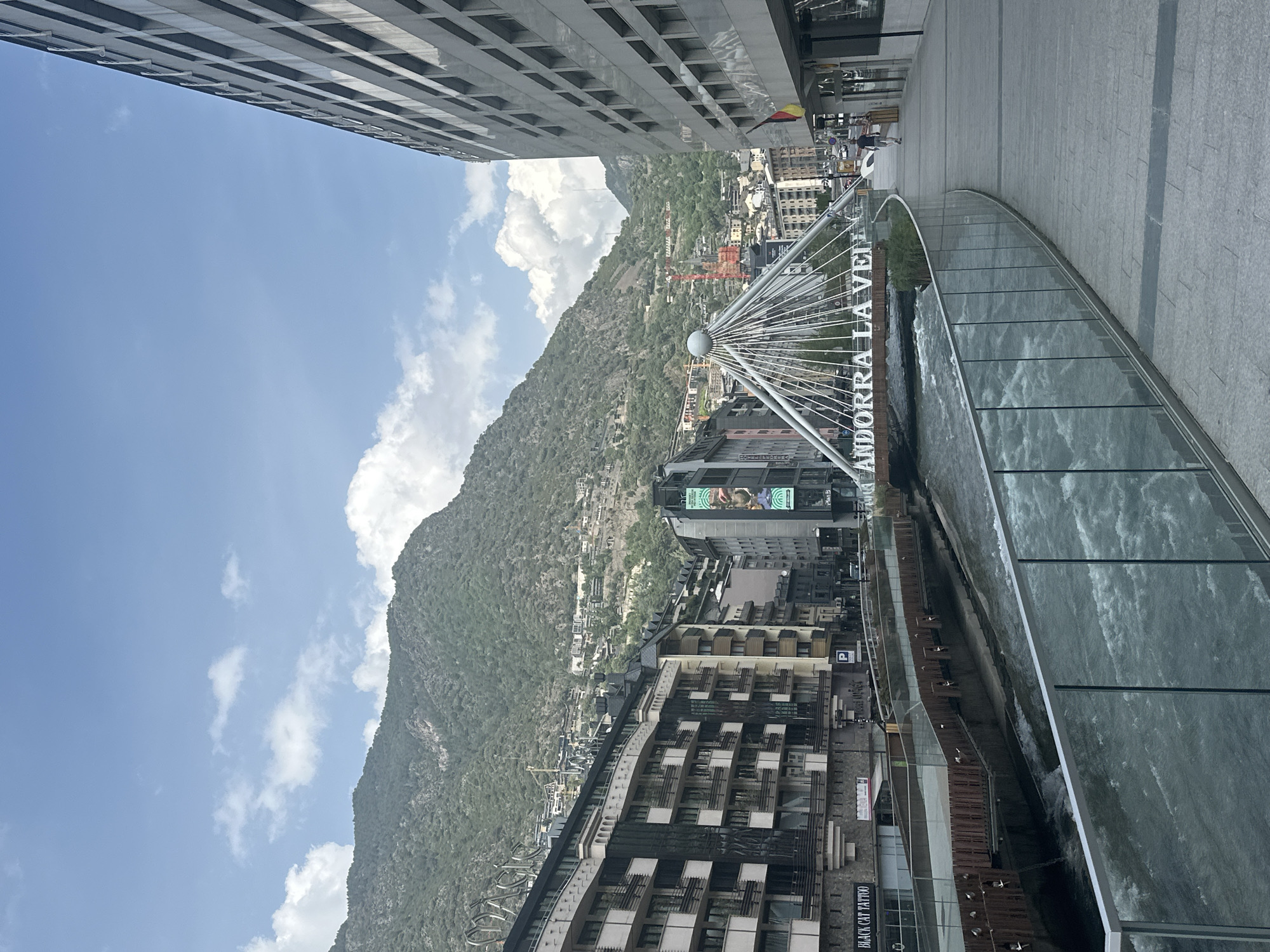 The image depicts a cityscape of Andorra la Vella, Andorra, with a focus on the "Andorra la Vella" bridge, which is a modern steel structure with white lettering. A river flows beneath the bridge, contained by glass barriers. A large mountain covered in greenery rises behind the urban development, with buildings interspersed across its lower slopes. In the foreground, the edge of a tall, modern building is visible. The sky is blue with scattered white clouds. People are visible walking along the elevated walkway.