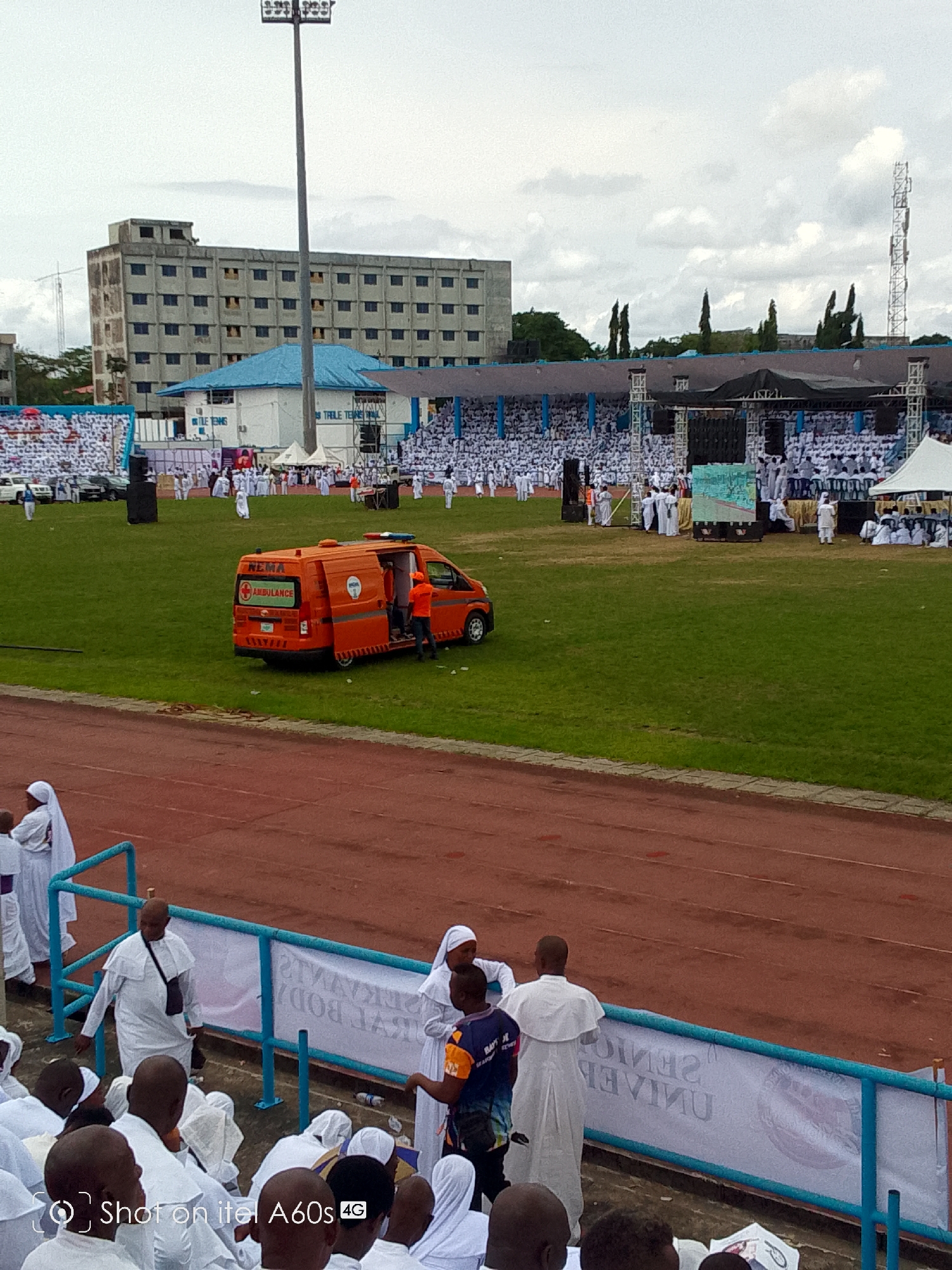 This image captures a large gathering of people, primarily dressed in white robes, at what appears to be a stadium or sports field in Calabar, Nigeria. The scene is viewed from an elevated position, looking down towards the field and the stands.
In the foreground, a red running track encircles a green grass field. Several individuals in white attire are visible near the track, some standing and some seated. A light blue metal railing separates the spectators from the track.
On the grass field, a prominent orange NEMA (National Emergency Management Agency) ambulance is parked, with its side door open and a person in an orange vest standing near it. Further back on the field, numerous people in white are gathered, along with a large stage structure with speakers and a screen. Several white tents are also visible on the field.
In the background, beyond the field and spectator stands, there's a large, multi-story concrete building with many windows, appearing somewhat aged. Next to it are smaller buildings with blue roofs. A tall stadium light tower stands prominently in the center-left, reaching towards the cloudy sky. To the right, a communication tower is visible.
The weather appears overcast, suggesting a cloudy day. The atmosphere indicates a large-scale public event, possibly religious or ceremonial, given the predominant white attire worn by most attendees.
Visible text includes "NEMA AMBULANCE" on the orange vehicle, and "SHOT ON ITEL A60S 4G" as a watermark in the bottom left. Partially visible and reversed text on banners near the track reads "CALABAR", "SERAPHIC", and "SENIOR UNIVERSITY". Another banner on a building in the background shows "BLE TENNIS".