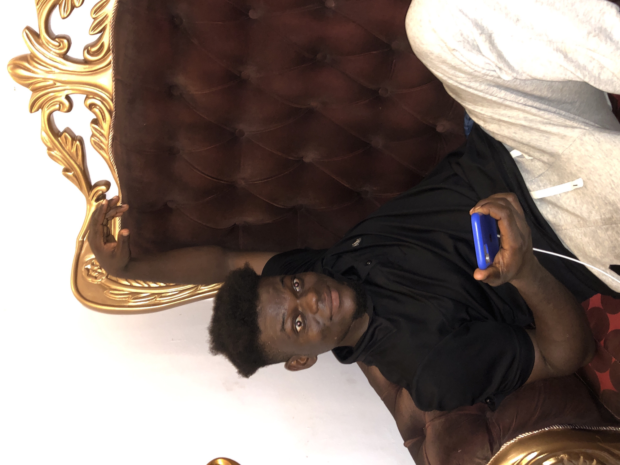 A person is lying on a tufted, dark brown couch with gold trim. The person has dark skin, short, curly black hair, and is wearing a black shirt. The person is holding a blue phone in their right hand. The couch is positioned against a plain white wall. A person wearing gray sweatpants is also sitting on the couch, partially visible on the upper right of the image. The location is Unknown, Unknown.