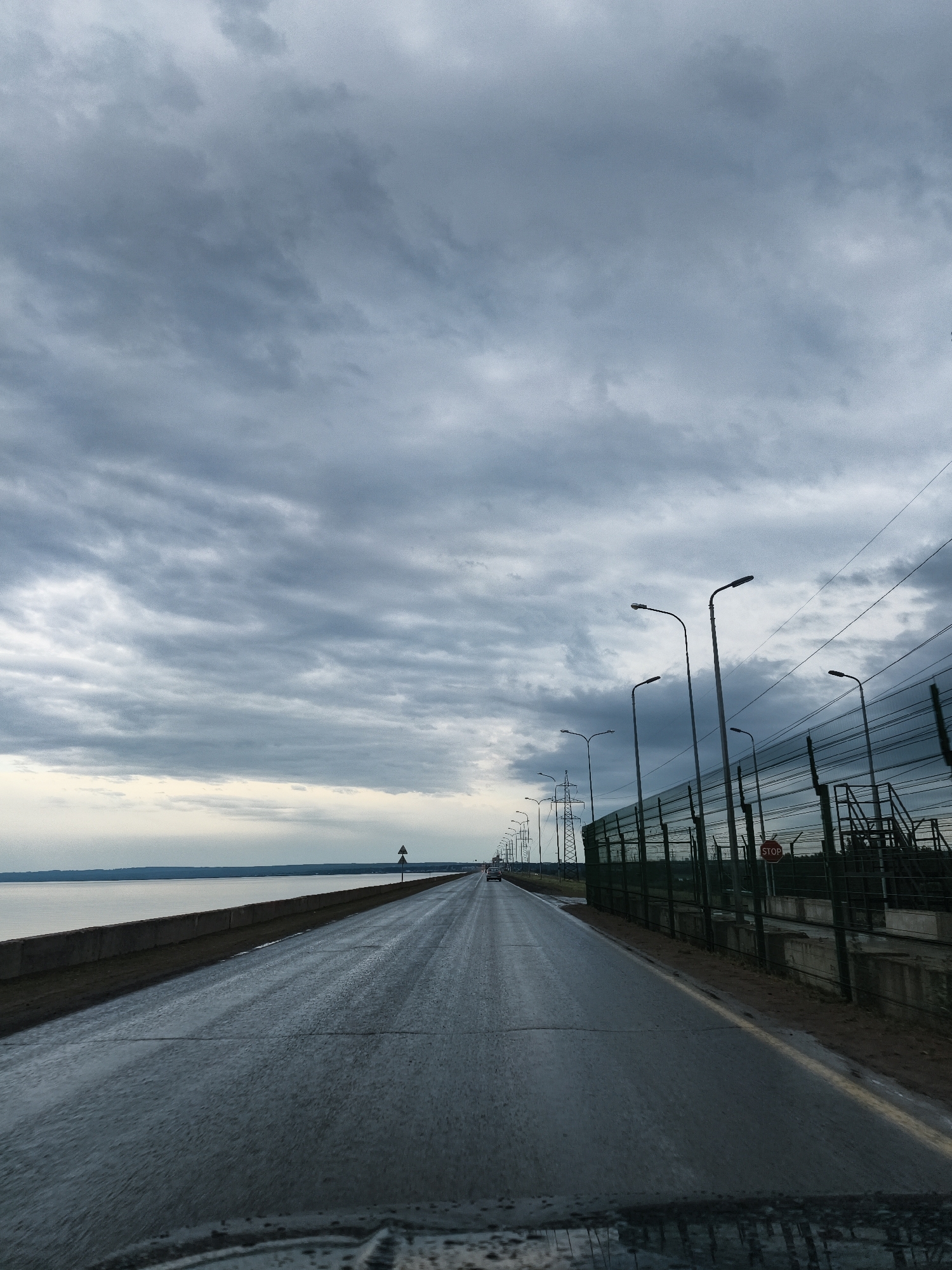 The image depicts a wet, asphalt road stretching into the distance under a cloudy, overcast sky. To the left of the road is a body of water, likely a river or lake, with a concrete embankment. The far shoreline is visible in the distance. To the right of the road is a tall metal fence with a sound barrier and a visible "STOP" sign attached. Street lights line the right side of the road alongside electrical pylons. The scene is taken from inside a vehicle, with a portion of the dashboard visible at the bottom. The location is Noviy, Russia. A vehicle is visible in the distance on the road.