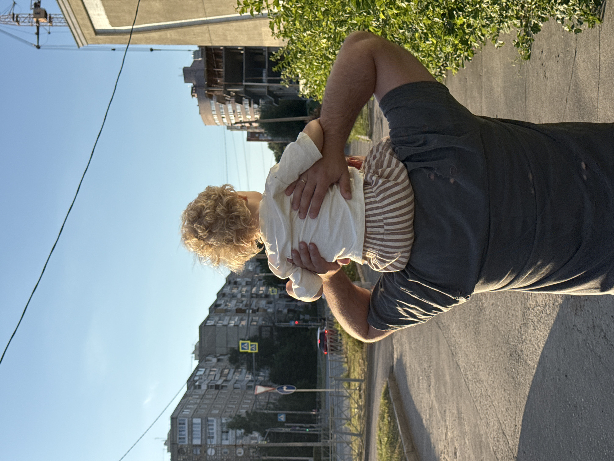 A person is holding a baby on their shoulders. The baby has blonde curly hair and is wearing a white shirt with striped sleeves underneath. The person is wearing a dark grey t-shirt. They are standing on a paved surface, possibly a sidewalk or road, with a shadow cast to the right. In the background, there are several apartment buildings with many windows. There are trees with green leaves visible. A street scene unfolds with traffic lights and road signs. The sky is clear and blue, suggesting it is daytime. A construction crane is visible in the upper left portion of the image, indicating potential ongoing construction. The overall scene appears to be in an urban environment. The lighting suggests it is late afternoon or early evening, with the sun casting long shadows. The context provided indicates the location is Vladimir, Russia. No text is clearly visible.