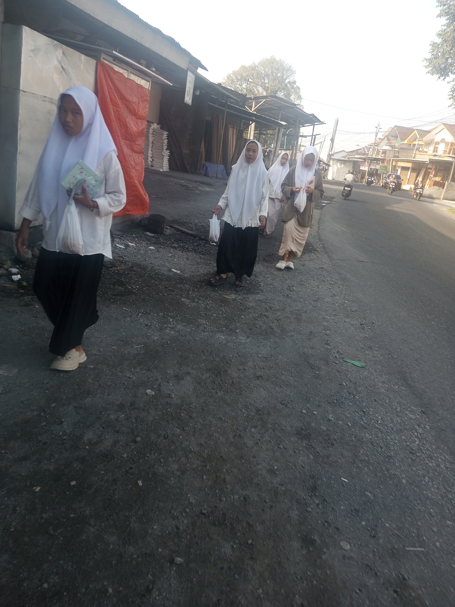 The image depicts a street scene in Bumirejo, Indonesia. Four young females, dressed in traditional Islamic attire with white hijabs and long dark skirts, are walking on the side of a paved road. The first girl on the left is holding a book and a white plastic bag. Behind her, two other girls walk closely, with the middle one holding a white plastic bag. A fourth girl is further behind, also in similar attire. To the left of the girls, there are structures that appear to be shops or stalls with corrugated metal roofs, one draped with an orange tarp. Stacks of wood and other materials are visible behind the stalls. The road itself is grey asphalt, with patches of loose gravel and debris on the shoulder. In the background, there are buildings and a few motorcycles are visible on the road, suggesting ongoing activity in the area. The sky is bright and overcast.