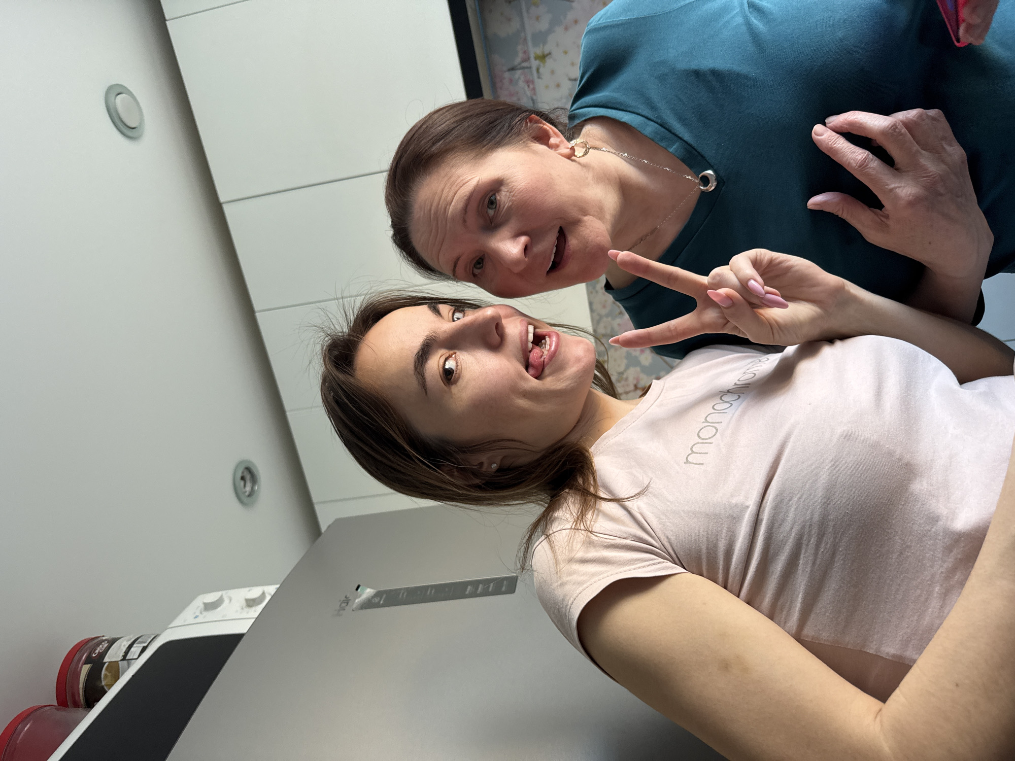 Two women are posing for a selfie indoors. One woman wears a teal shirt and a necklace, and the other wears a pink shirt and is making a peace sign while sticking out her tongue. A refrigerator is visible in the foreground. Cans or jars are to the left of the refrigerator. The location could potentially be Sarapul, Russia, but there are no verifiable elements in the image to confirm this. The environment appears to be a kitchen or eating area.