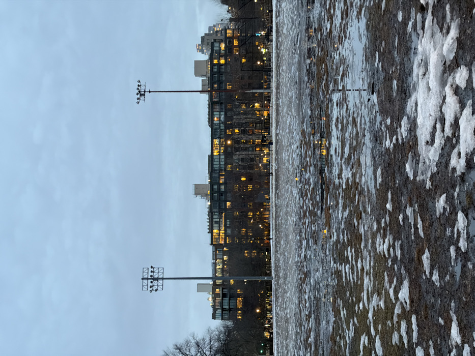 The photo shows a cityscape at dusk, with a field in the foreground partially covered in melting snow. Two tall light posts are visible on the left, with buildings behind them. The buildings are lit, casting a warm glow. The sky is overcast. The field is muddy and wet, with patches of white snow. The image is slightly tilted.