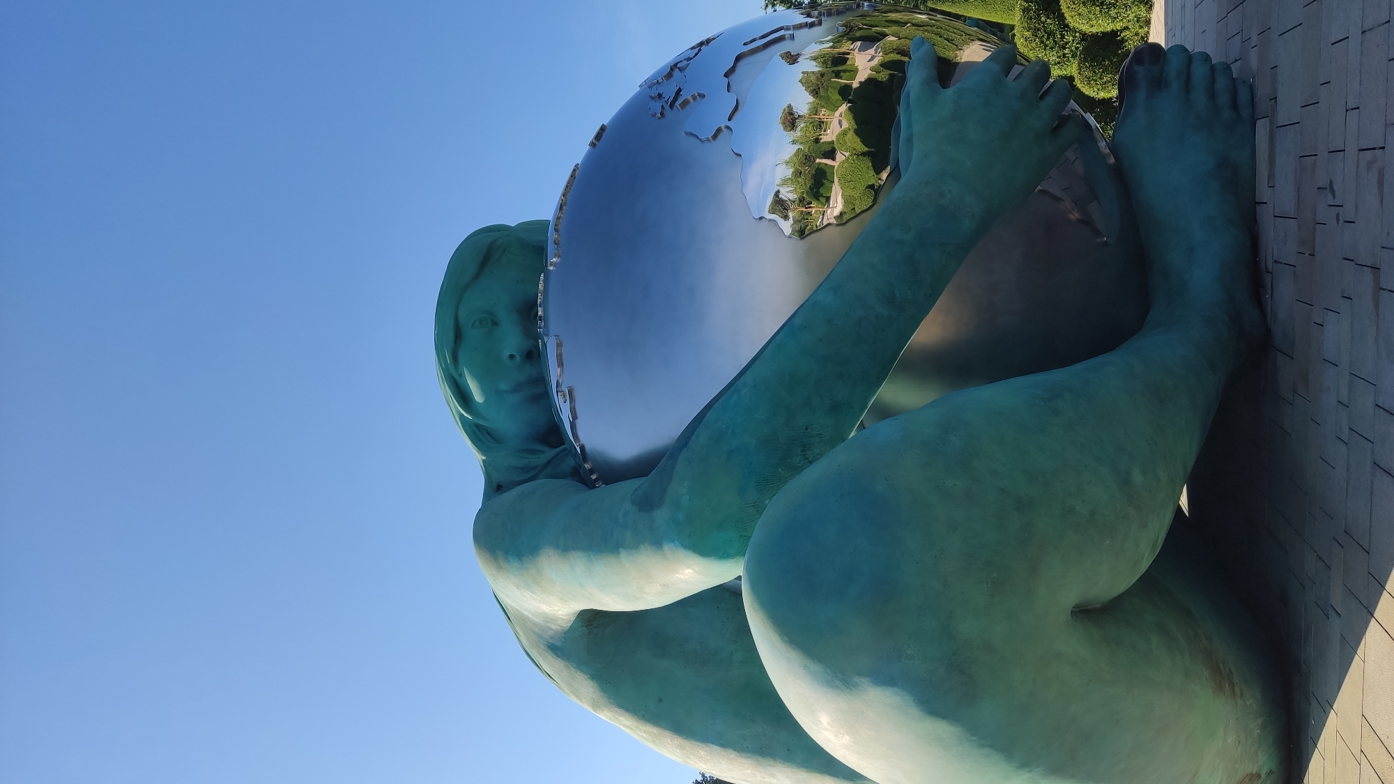 The image shows a large, teal-colored sculpture of a person embracing a metallic sphere that resembles the Earth. The sculpture is positioned outdoors, with a clear blue sky in the background and a paved ground with a brick pattern. The metallic sphere reflects the surrounding environment, showing trees, bushes, and possibly buildings. The sculpture's posture suggests a protective or intimate embrace of the Earth. This sculpture is located in the city of Krasnodar, Russia.
