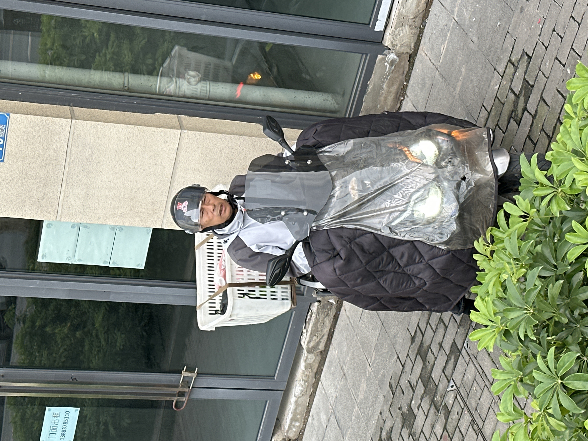 A man wearing a helmet and jacket sits on a scooter parked next to a building in the city of Jiangjin, China. The scooter has a plastic sheet covering the front. A white basket is attached to the front of the scooter. The building has large windows, with green vegetation visible outside and a sign in Chinese characters posted on the door. The sidewalk is paved with grey brick.