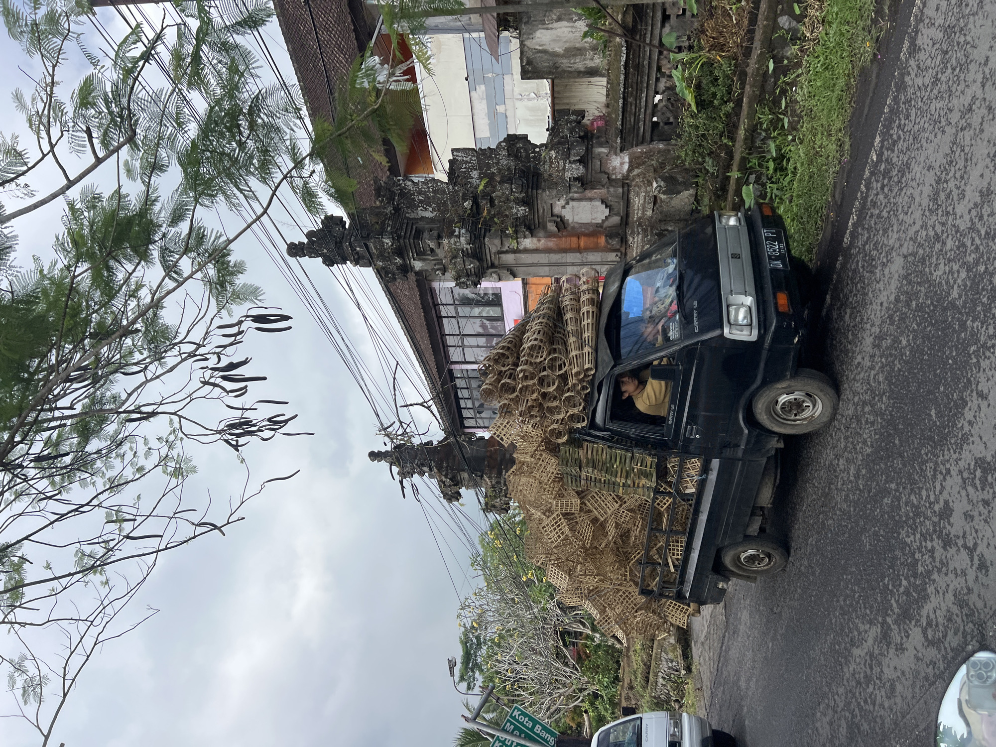A black Suzuki Carry pickup truck is driving on an asphalt road in Tembuku, Indonesia. The truck's bed is heavily loaded with woven bamboo baskets. A man is visible driving the truck. In the background, there is a traditional Balinese gate and a building with a tiled roof. Overhead power lines are strung across the scene. The sky is overcast, and there are trees with foliage and seed pods on the left side of the image. A white van is parked on the left side of the road. A green sign with white text is also visible on the left. The license plate on the truck reads "DK 8522 PT".