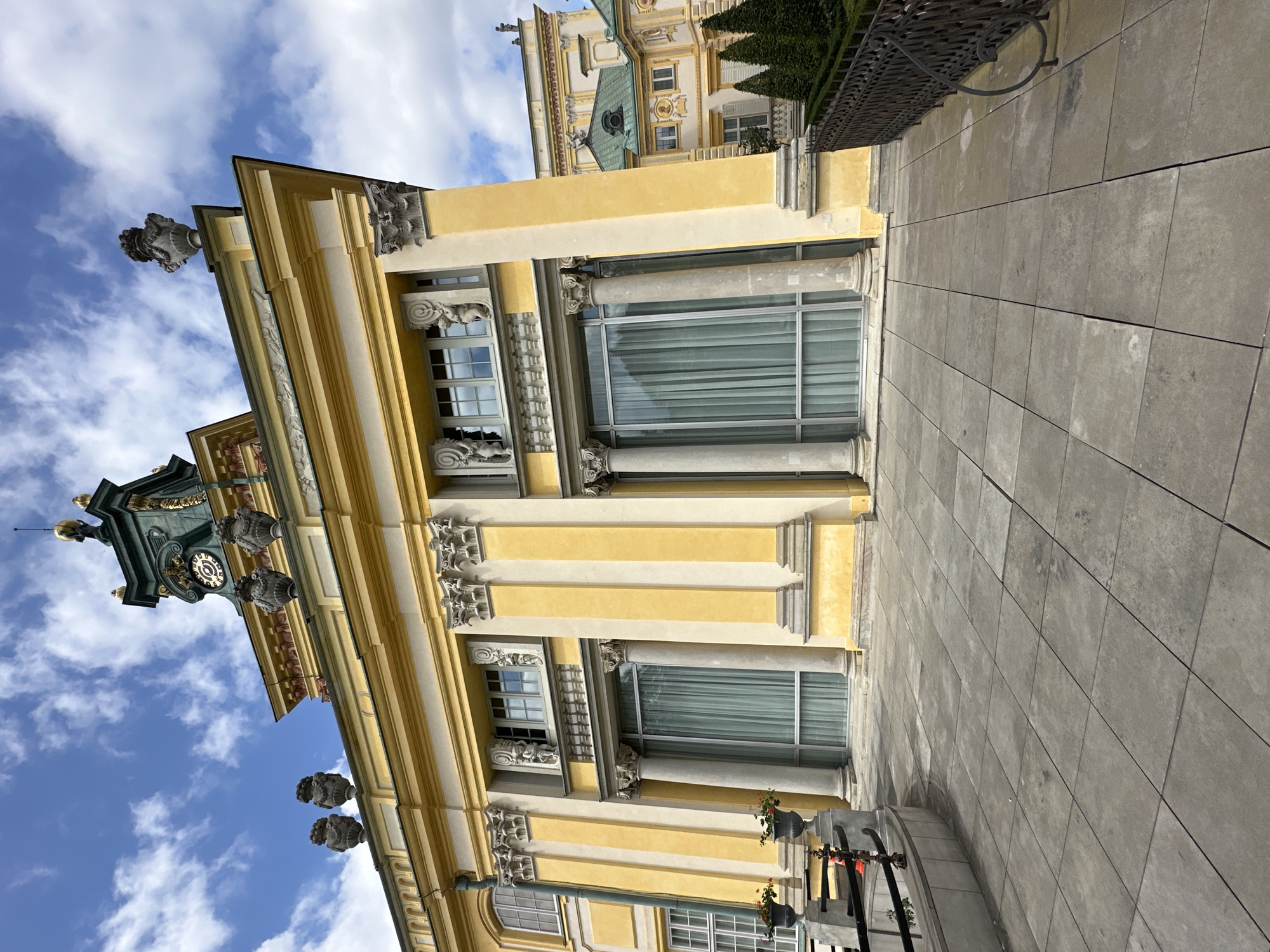The image shows the facade of the Wilanów Palace in Warsaw, Poland. The palace is a historic royal residence with a yellow facade, white columns, and ornate decorations. The sky is blue with scattered clouds. In the foreground, there is a paved courtyard with large stone tiles. In the background, there is a garden with manicured hedges and trees. The image is taken from a low angle, looking up at the palace. There are no people visible in the image.