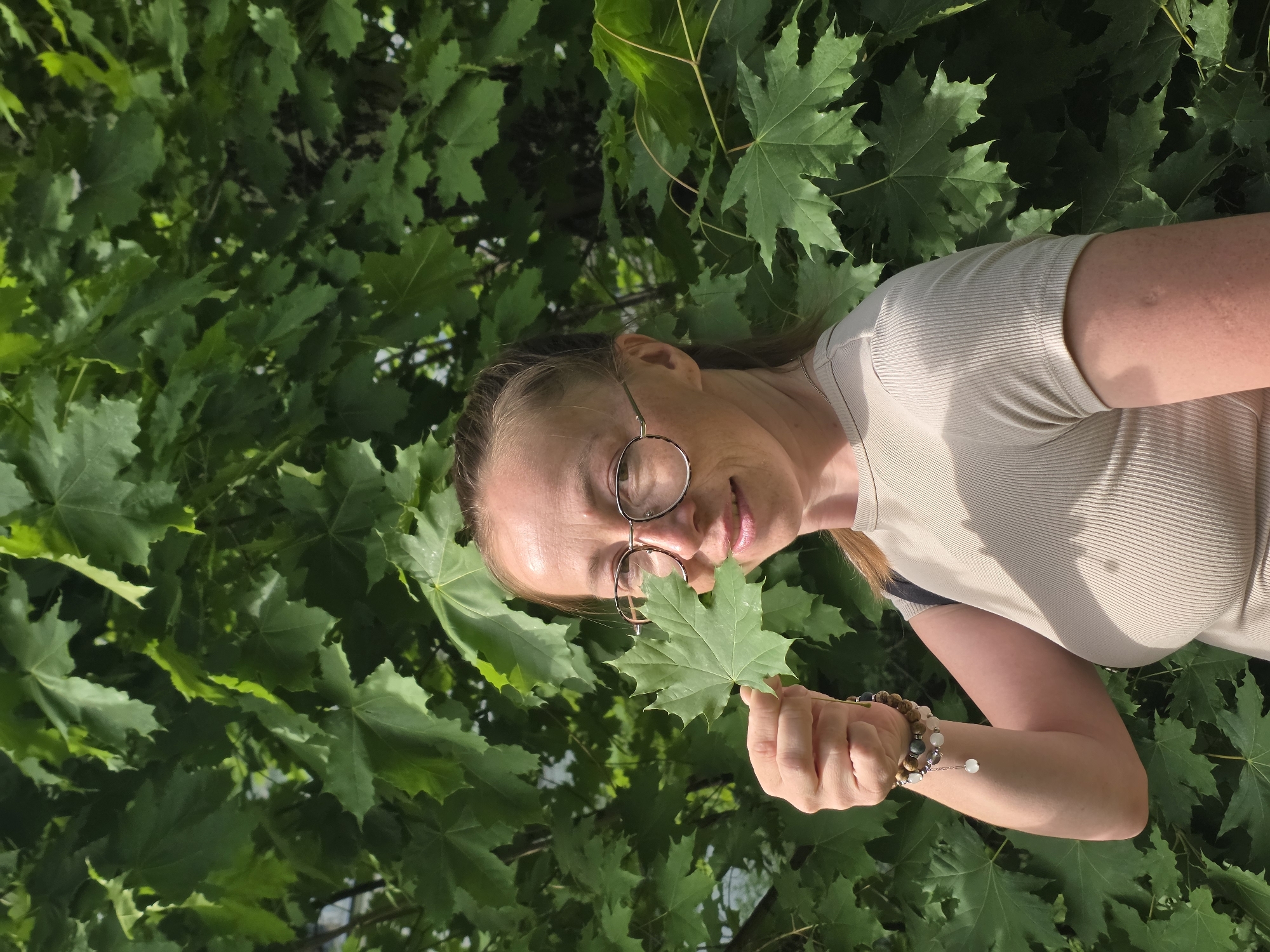 A woman wearing glasses and a bracelet stands outdoors in Chaikovsky, Russia, holding a green maple leaf in front of her face. The background consists of dense maple foliage. The woman is wearing a light-colored ribbed shirt.