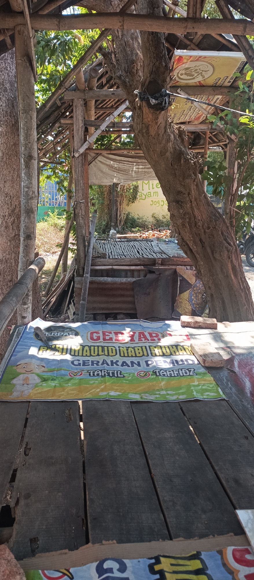 The image depicts an outdoor scene, likely in Jatisari, Indonesia, under a rustic, open-air shelter constructed from wood and bamboo. A large, weathered tree trunk dominates the center-right of the frame, its branches supporting parts of the shelter's roof. In the foreground, a wooden table is covered by a banner advertising a "GEBYAR MAULID NABI MUHAMMAD" event, which translates to "Maulid Nabi Muhammad Festival." The banner features illustrations of children in traditional Islamic attire and text mentioning "GERAKAN PEMUDA," "TARTIL," and "TEHFIDZ," suggesting a youth movement focused on religious activities.
Behind the shelter, a wall with "Mie ayam JOGJA" is visible, indicating a food stall or eatery. The environment appears somewhat unkempt, with vegetation growing around the structure and various materials like corrugated metal and tarpaulin used in its construction. Sunlight filters through the gaps in the shelter, creating patterns of light and shadow. The overall impression is of a simple, community gathering space, possibly associated with local religious celebrations and food vendors.