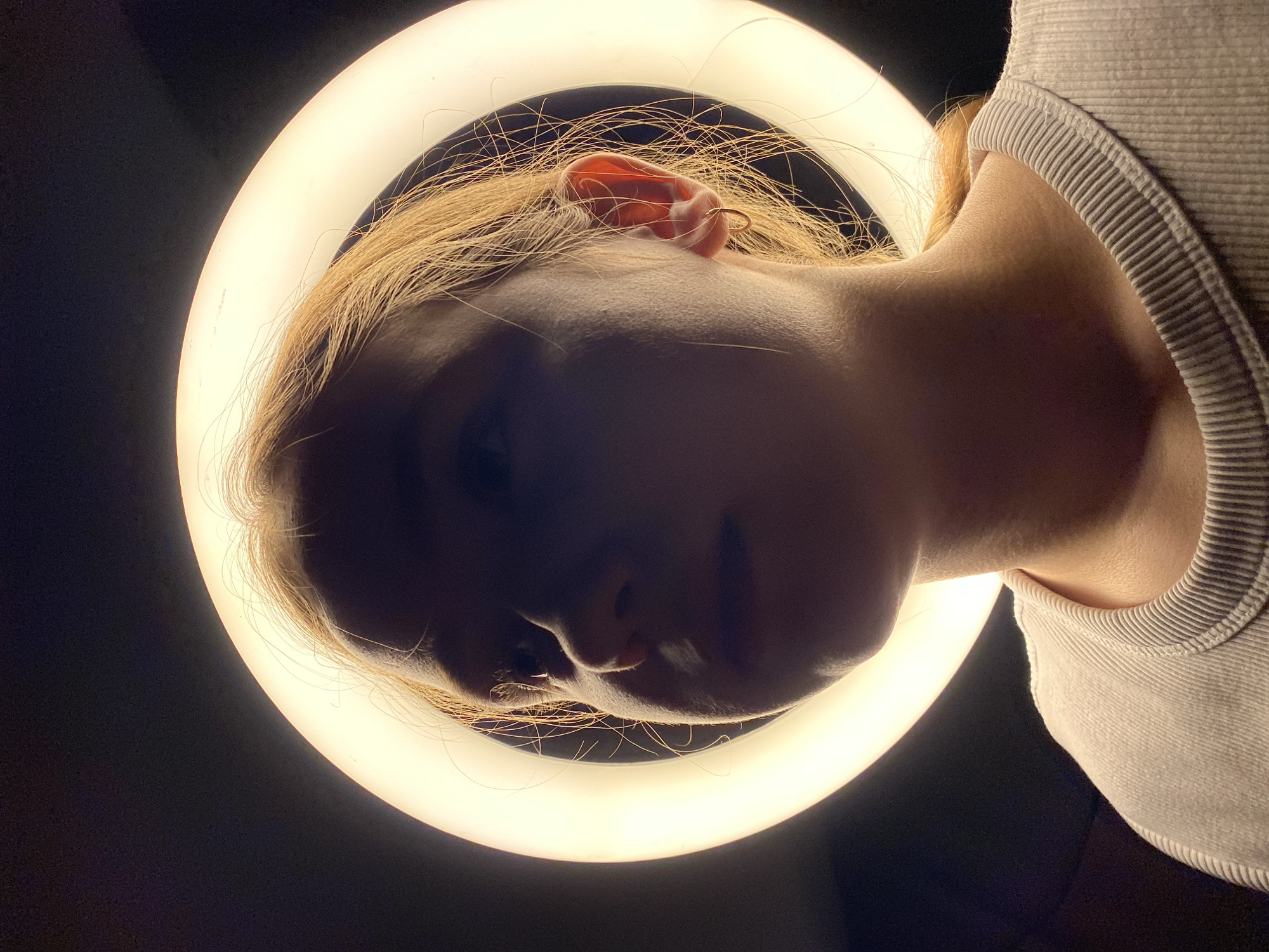 A low-angle shot shows a person’s face surrounded by a circular light. The person has fair skin and light brown or blonde hair. Their head is tilted, and their features are partially obscured by shadow. The circular light creates a halo effect around their head, and the background is dark. A portion of their shoulder and the collar of a light-colored shirt are visible on the right side of the frame. The location is Unknown, Unknown.