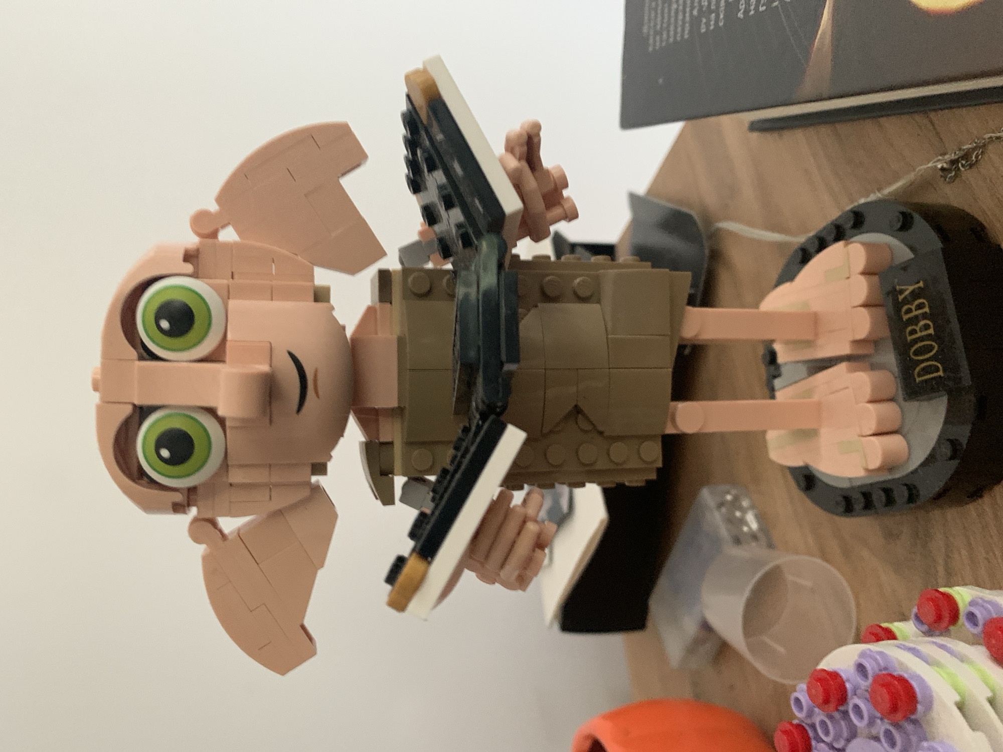 An eye-level, medium shot captures a Lego Dobby figurine standing on a wooden surface, likely a table. The figurine is composed of tan and brown Lego bricks, with large, green eyes and distinct tan ears. Dobby is holding two small, rectangular objects, each composed of white, black, and gold Lego pieces, in its hands. The background is a plain white wall. To the left of the figurine, a portion of an orange object and a clear plastic cup are visible. To the right, a book with a dark cover and some text is partially in frame. The base of the figurine has the word "DOBBY" printed on it in gold lettering. No specific location in Poznan, Poland can be determined from the image.